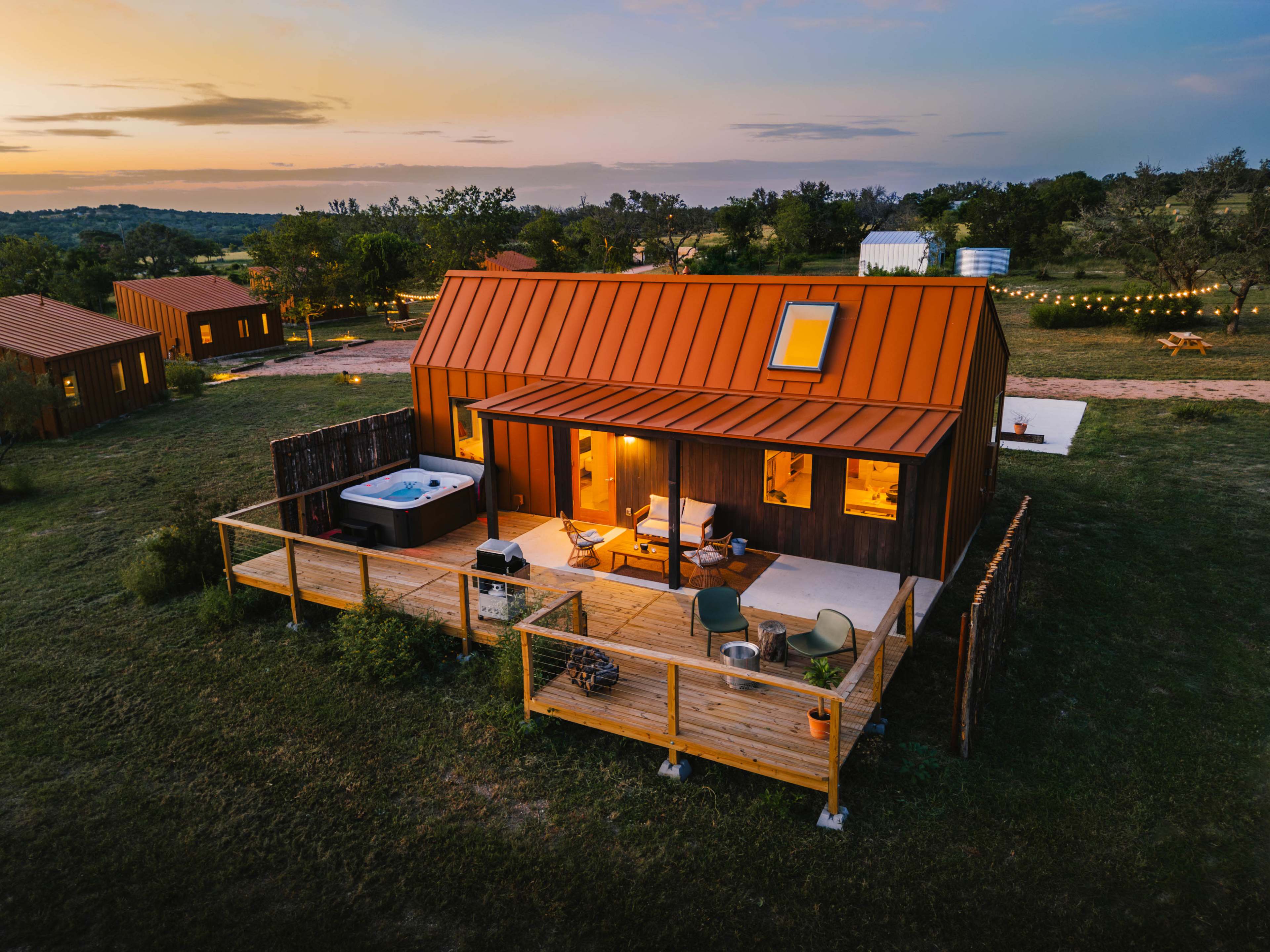 A modern cabin with a metal roof and wooden deck is illuminated at sunset, surrounded by green fields and a few other structures in the distance.
