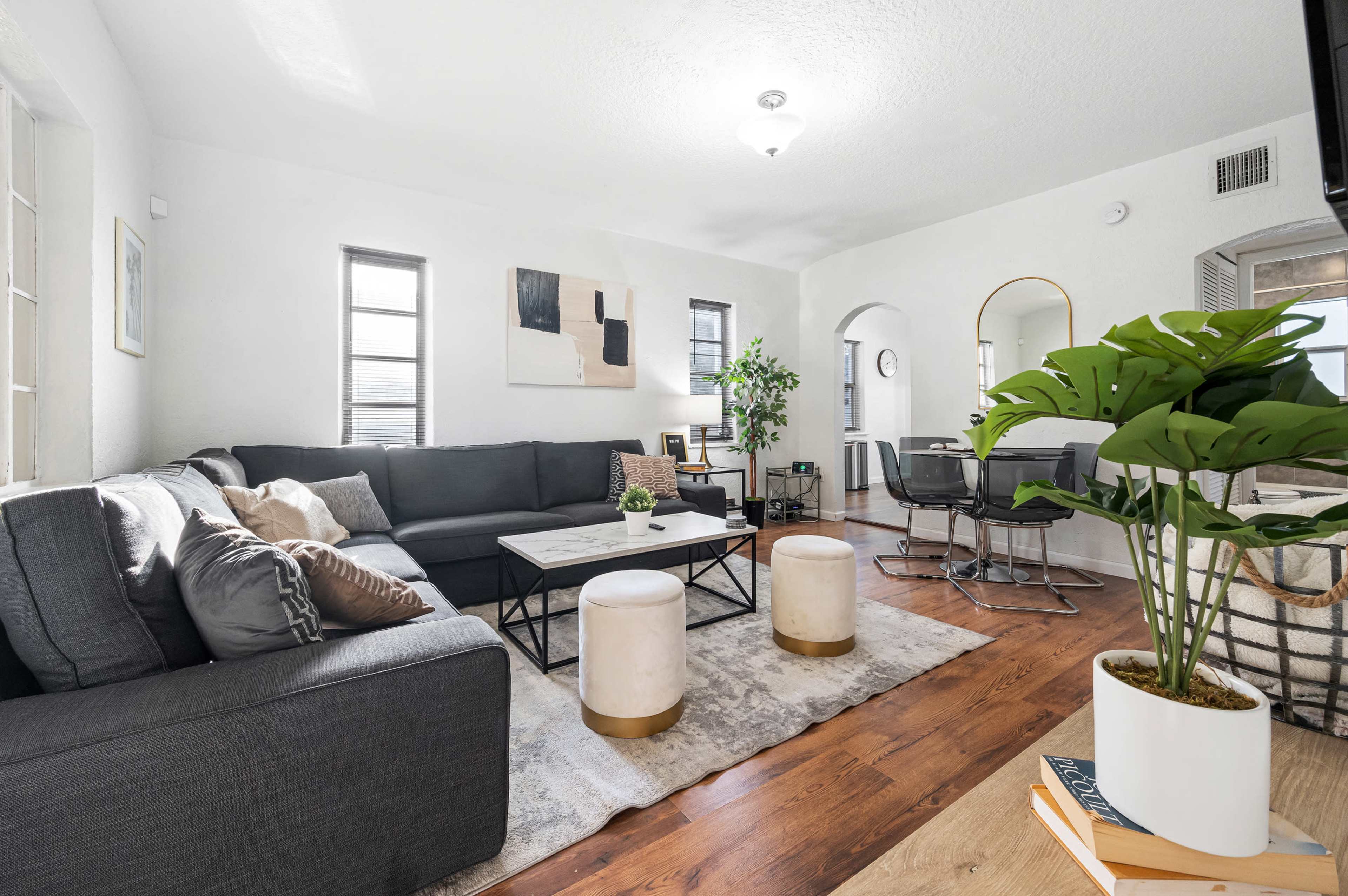 The living room features a gray sectional sofa, a coffee table with a decorative centerpiece, two ottomans, and a plant, all set against white walls and wooden flooring.