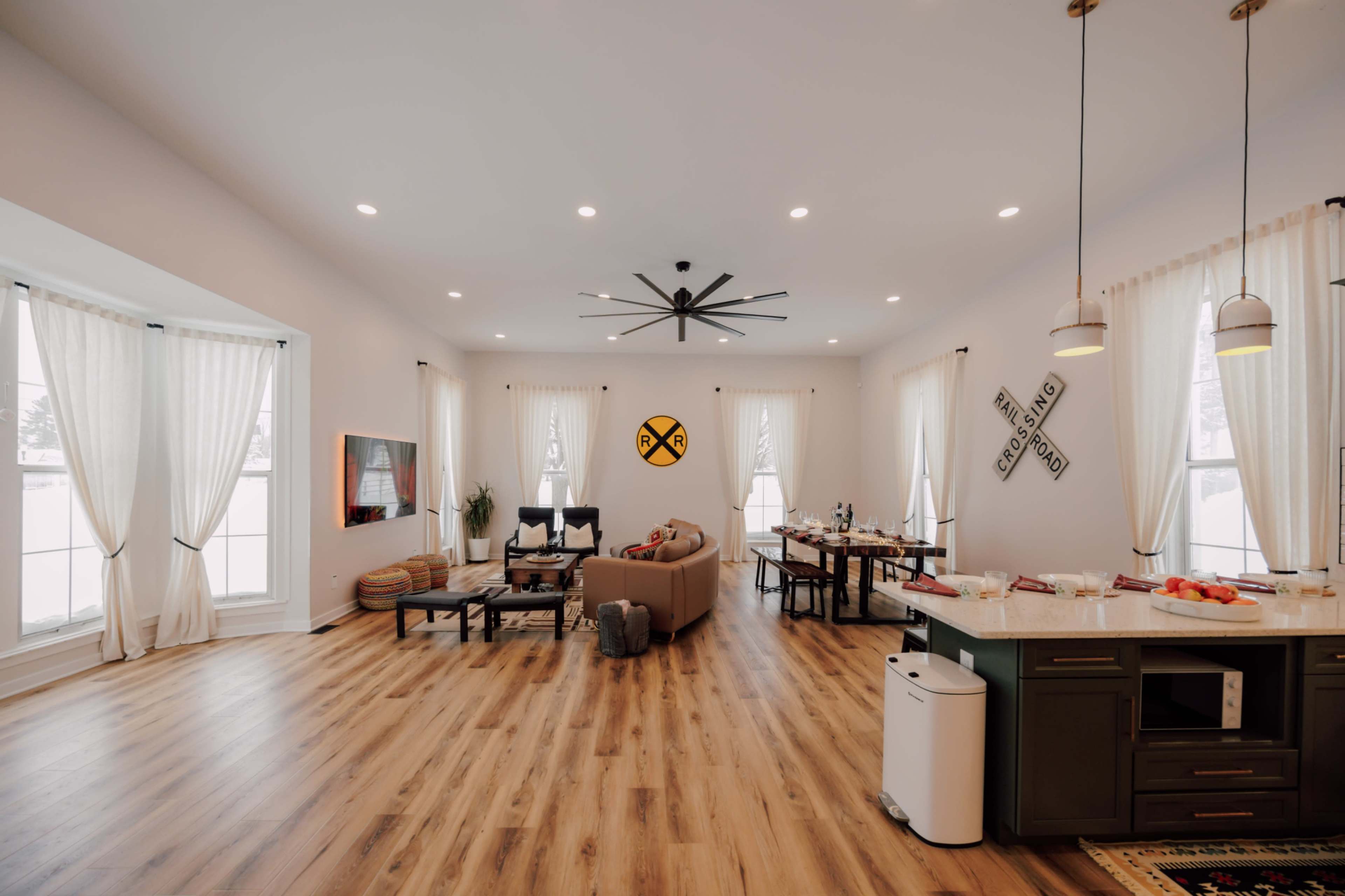An open living space features a sofa, dining area, and kitchen with modern fixtures, illuminated by natural light from large windows.