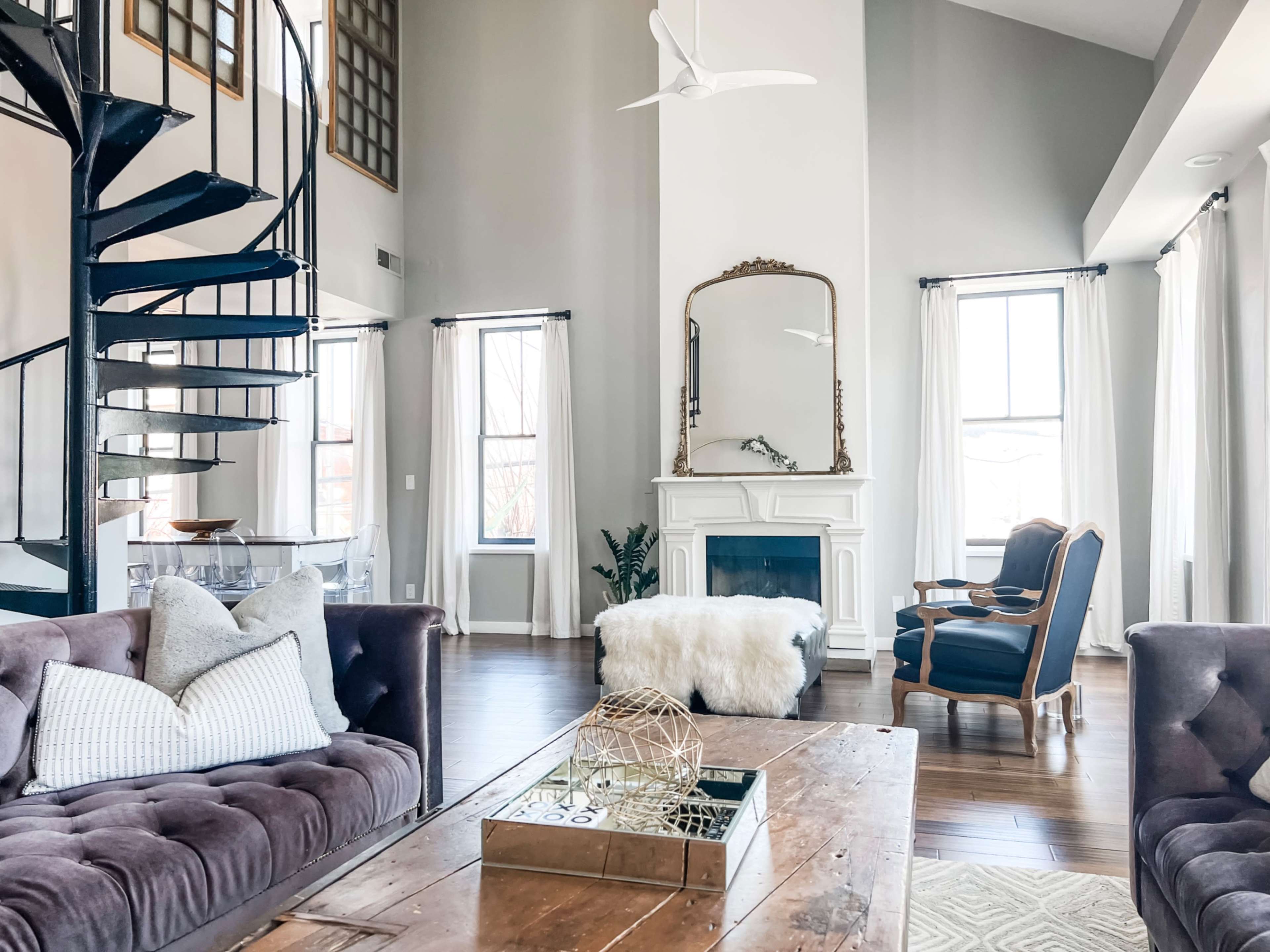 The image shows a spacious living room with a spiral staircase leading to an upper level, featuring a cozy seating area arranged around a rustic wooden coffee table and a white fireplace.