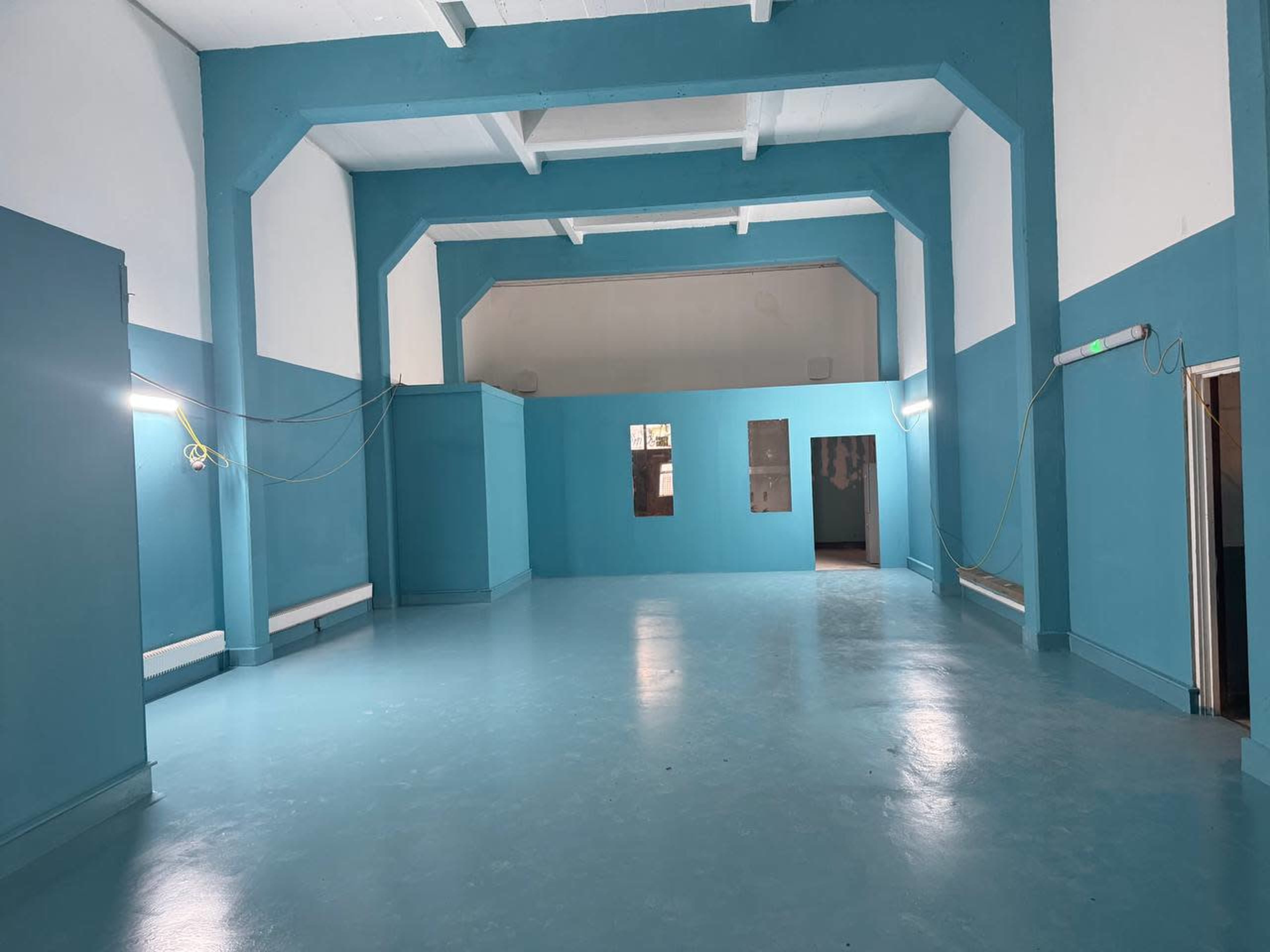The image shows a large, empty room with blue walls and a polished floor, featuring several light fixtures and doorways.