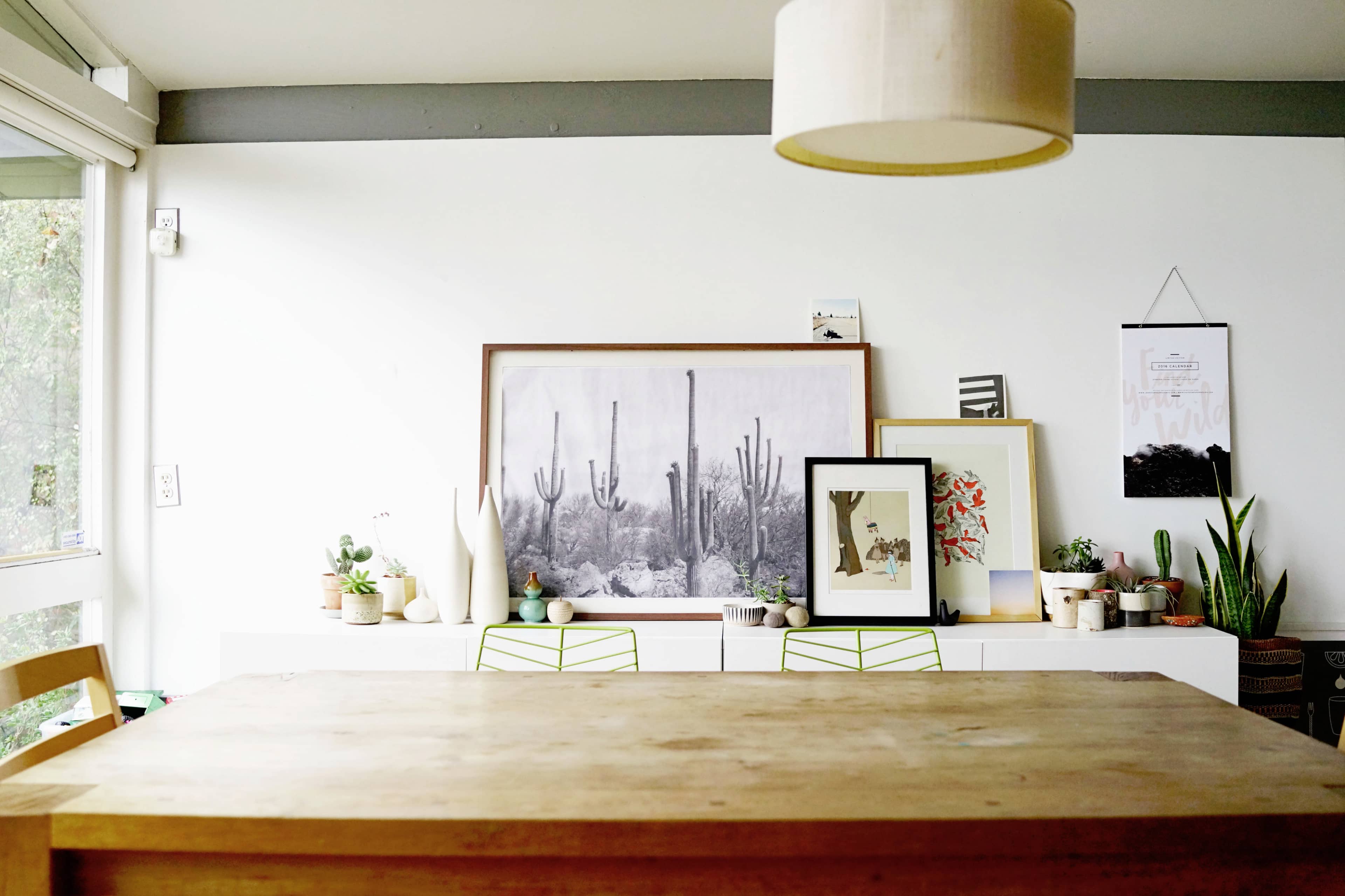 A wooden dining table is positioned in front of a gallery wall decorated with framed artworks and various potted plants, under a pendant light fixture.