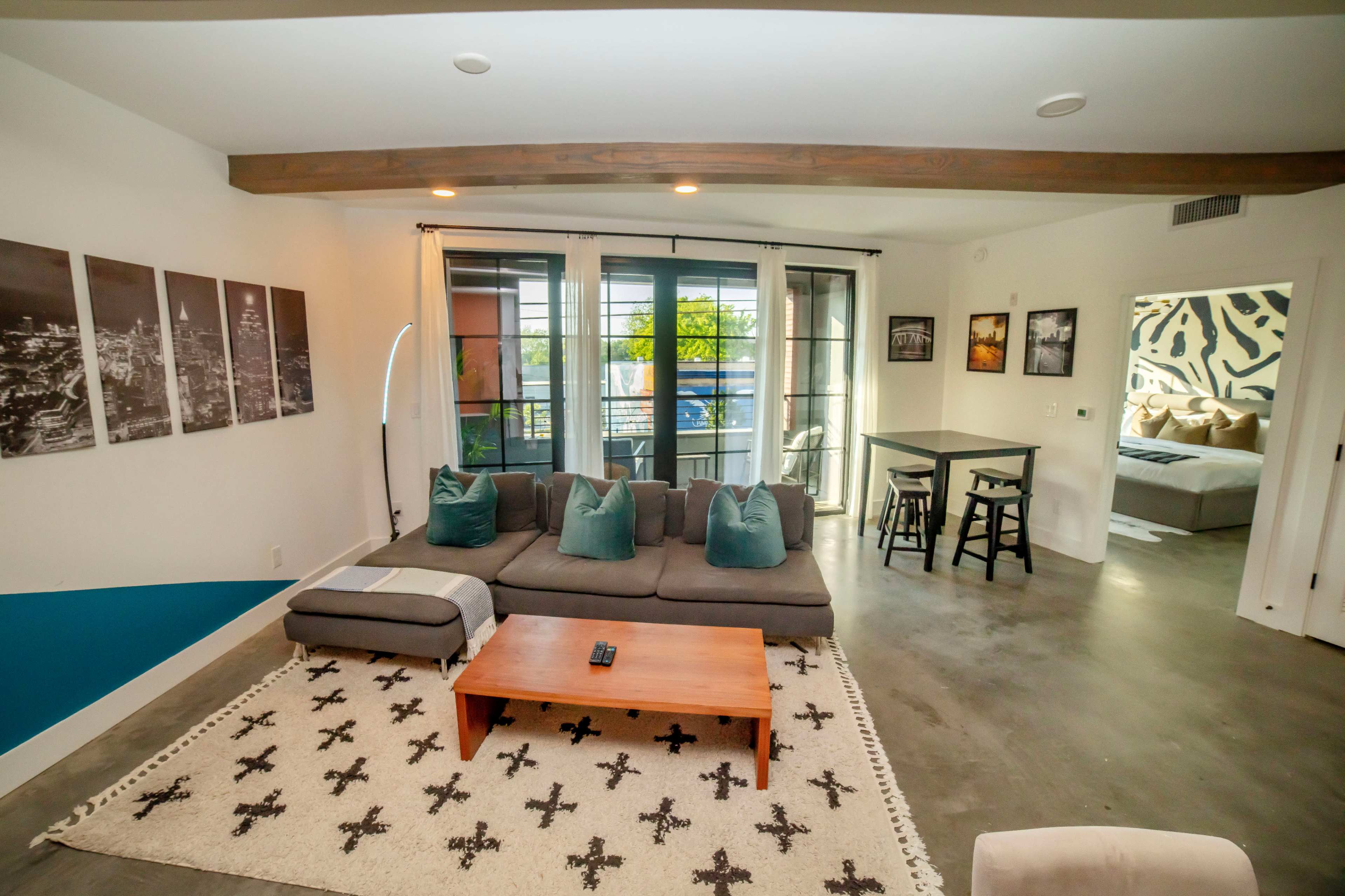 The image shows a modern living room with a sectional sofa, a coffee table, and a dining area in the background, featuring large windows and decorated walls.