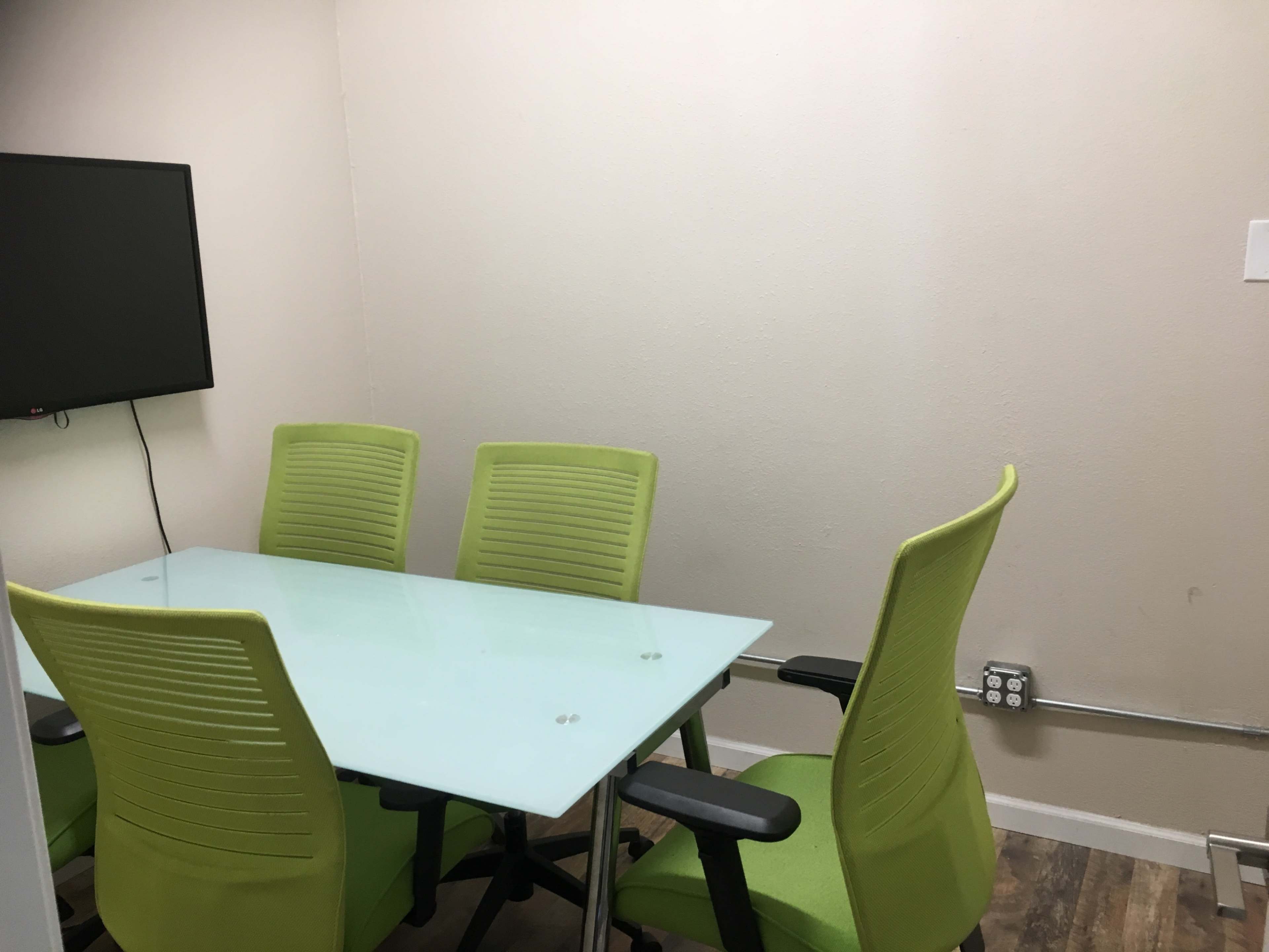A small meeting room features a glass table surrounded by four green chairs and a wall-mounted television.