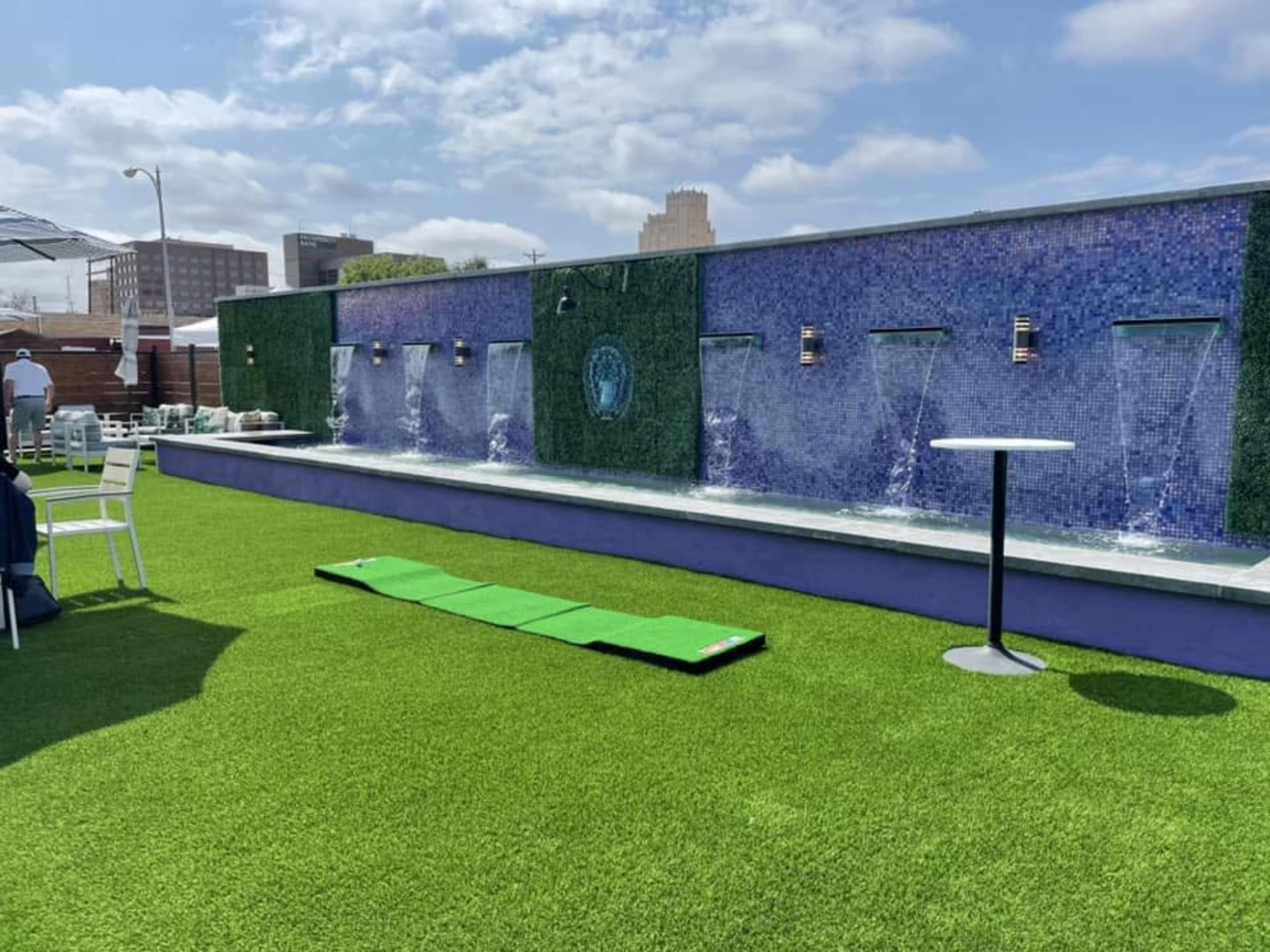 The image shows a rooftop area featuring a green artificial turf surface, a wall with multiple water fountains, and a small table beside a pathway.