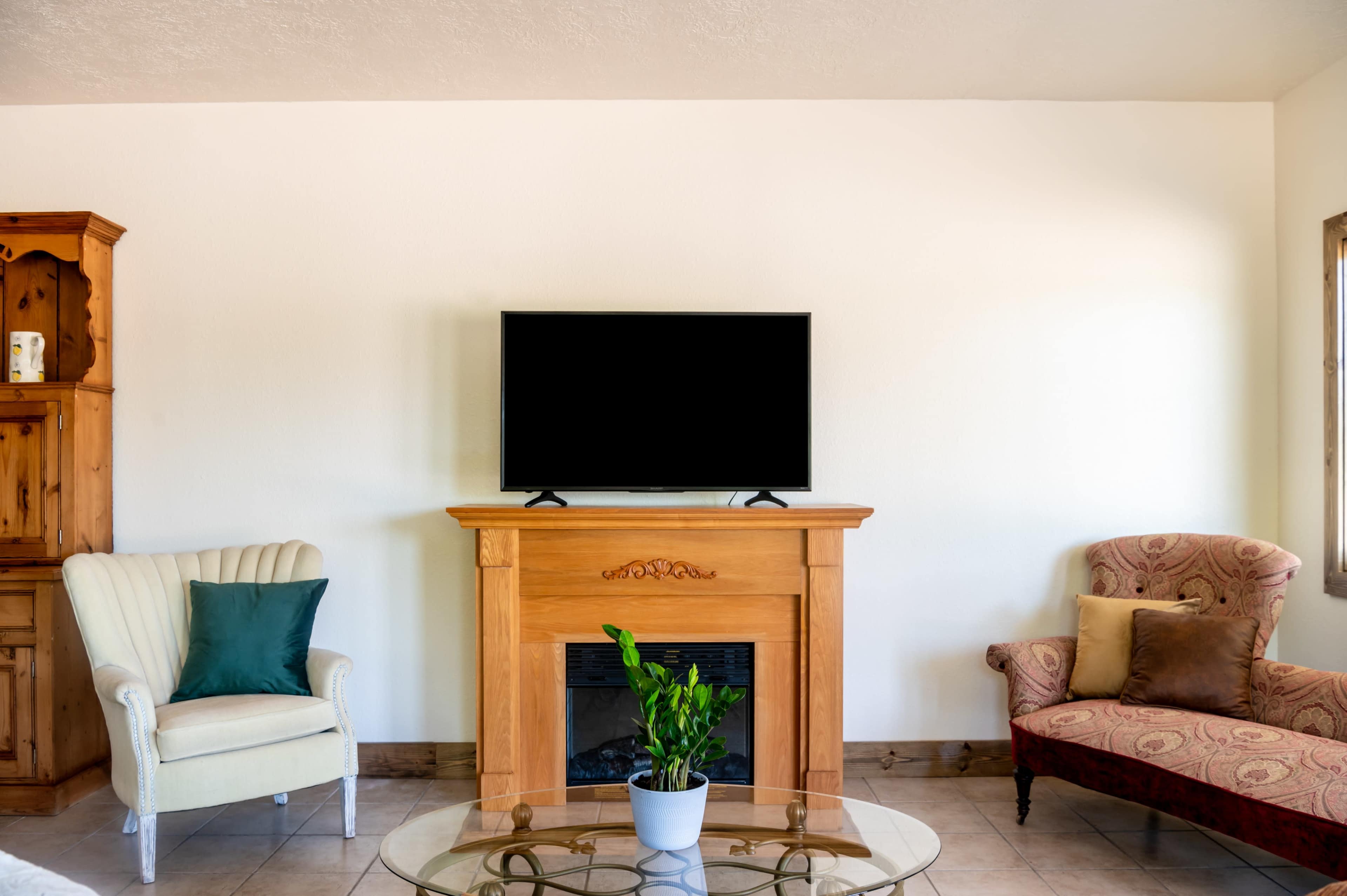 A living room with a wooden fireplace, a television mounted above it, and two chairs arranged around a glass coffee table.