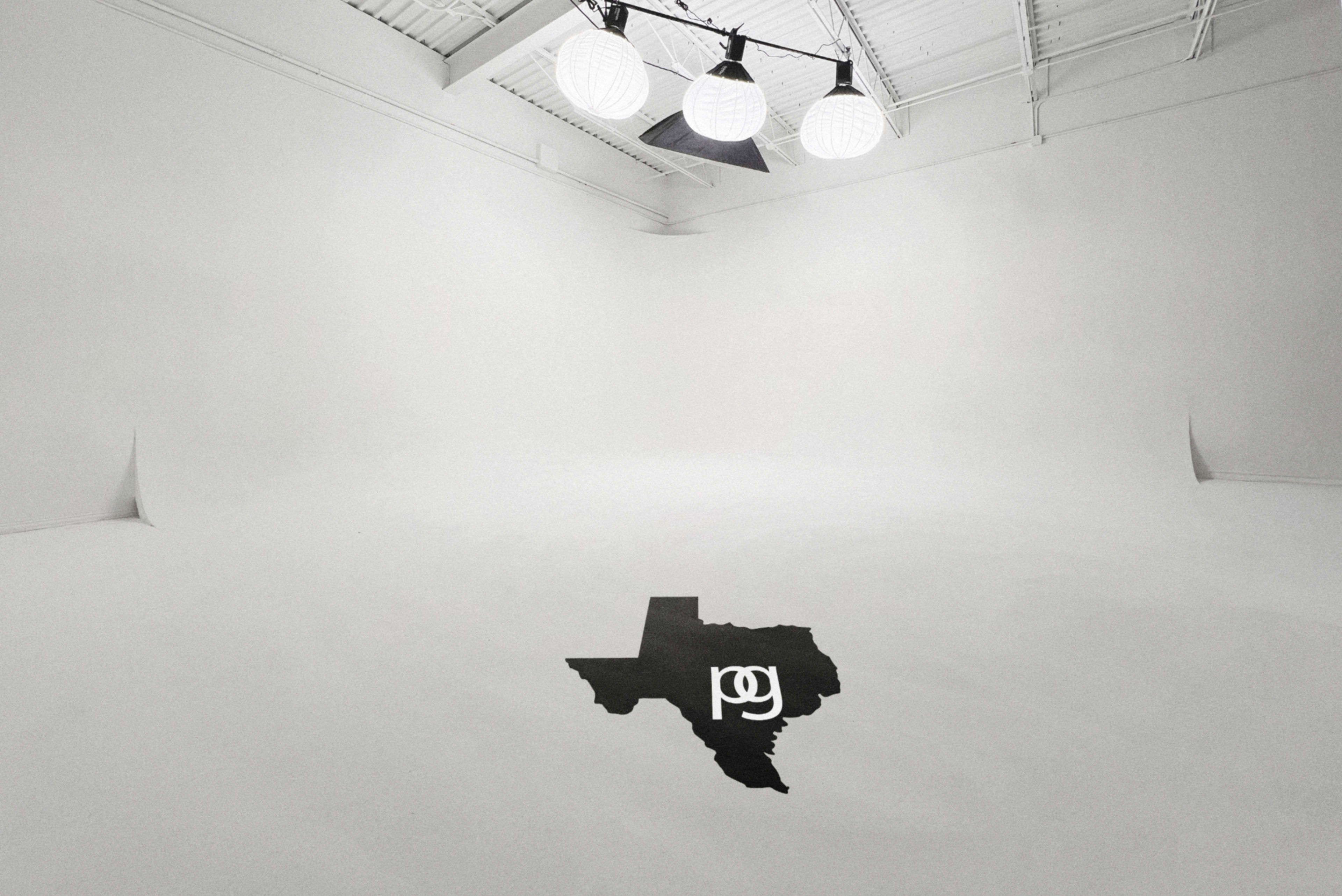 A spacious, empty studio with white walls and ceiling features three large round lights hanging above a black outline of the state of Texas on the floor.