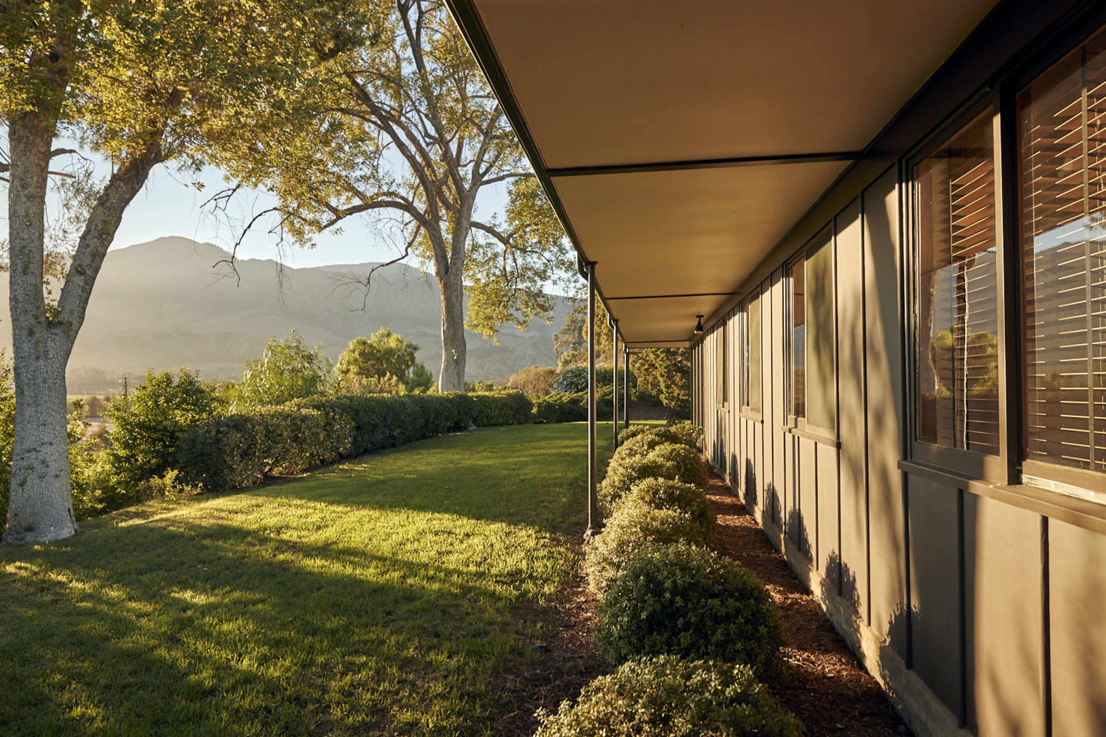 The image shows a long building with windows, bordered by well-maintained hedges and a grassy lawn, set against a backdrop of mountains and trees.
