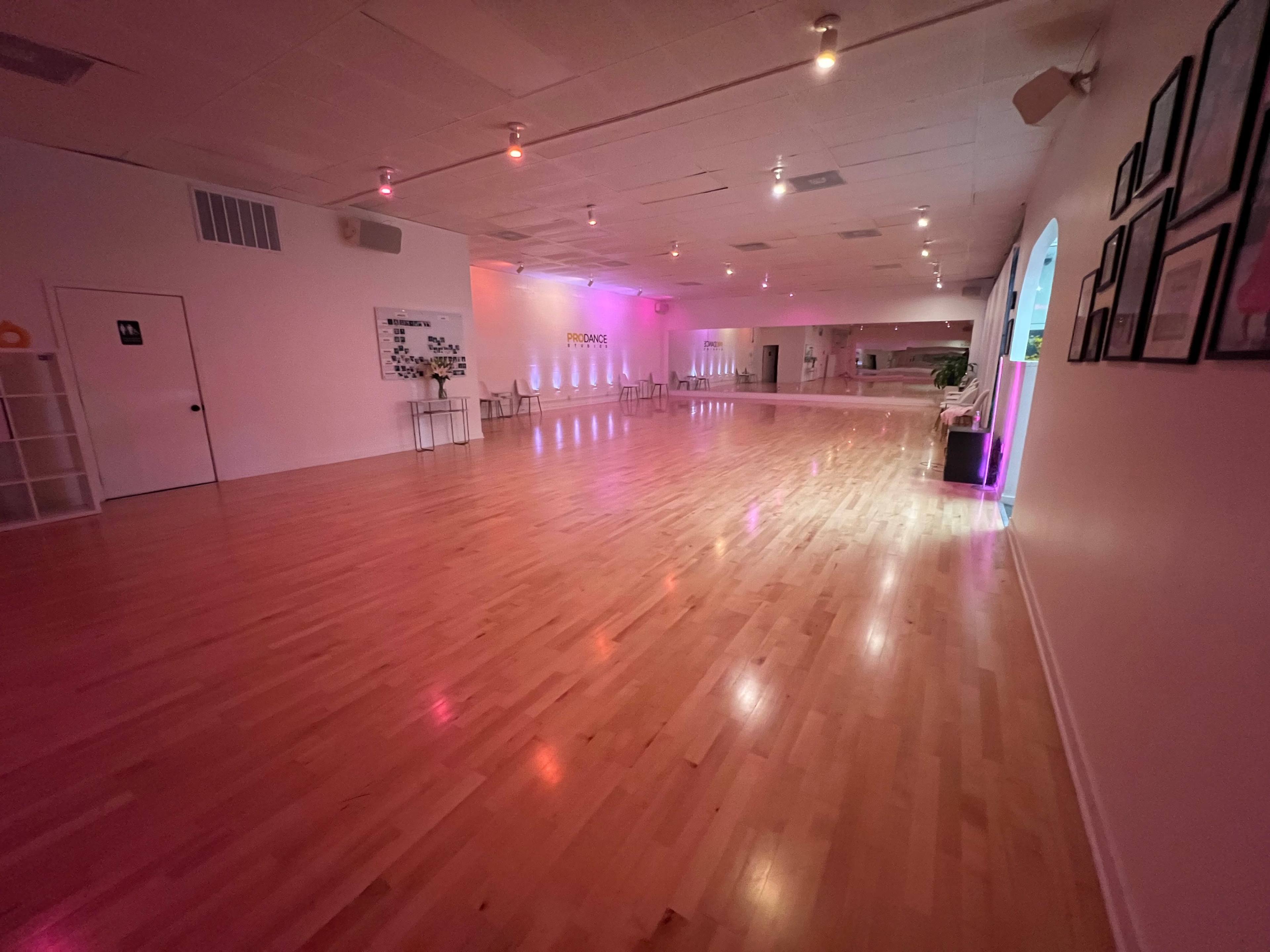 The image shows a spacious dance studio with polished wooden floors and soft, colorful lighting.