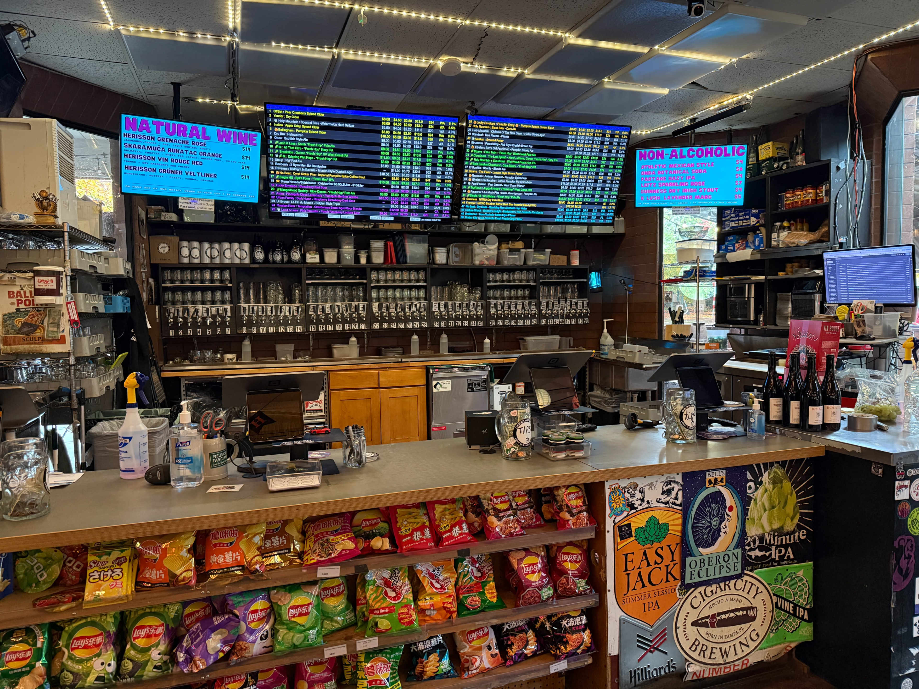 The image shows a bar counter with multiple taps, extensive menu boards displaying drink options, and a shelf filled with snacks and bottled beverages.