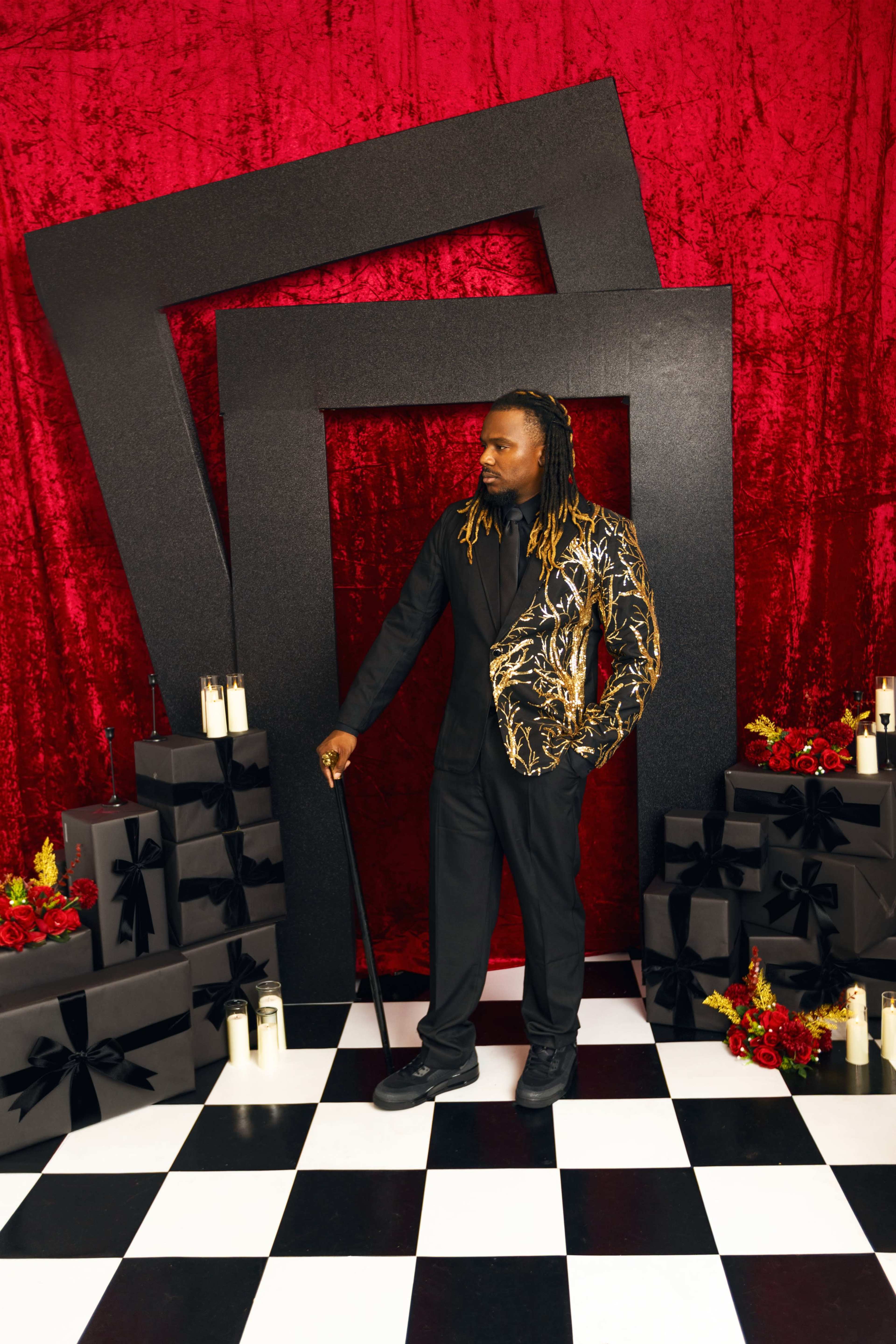 A man stands confidently in front of a striking red backdrop, framed by large black geometric shapes, surrounded by gifts adorned with black ribbons and lit candles.