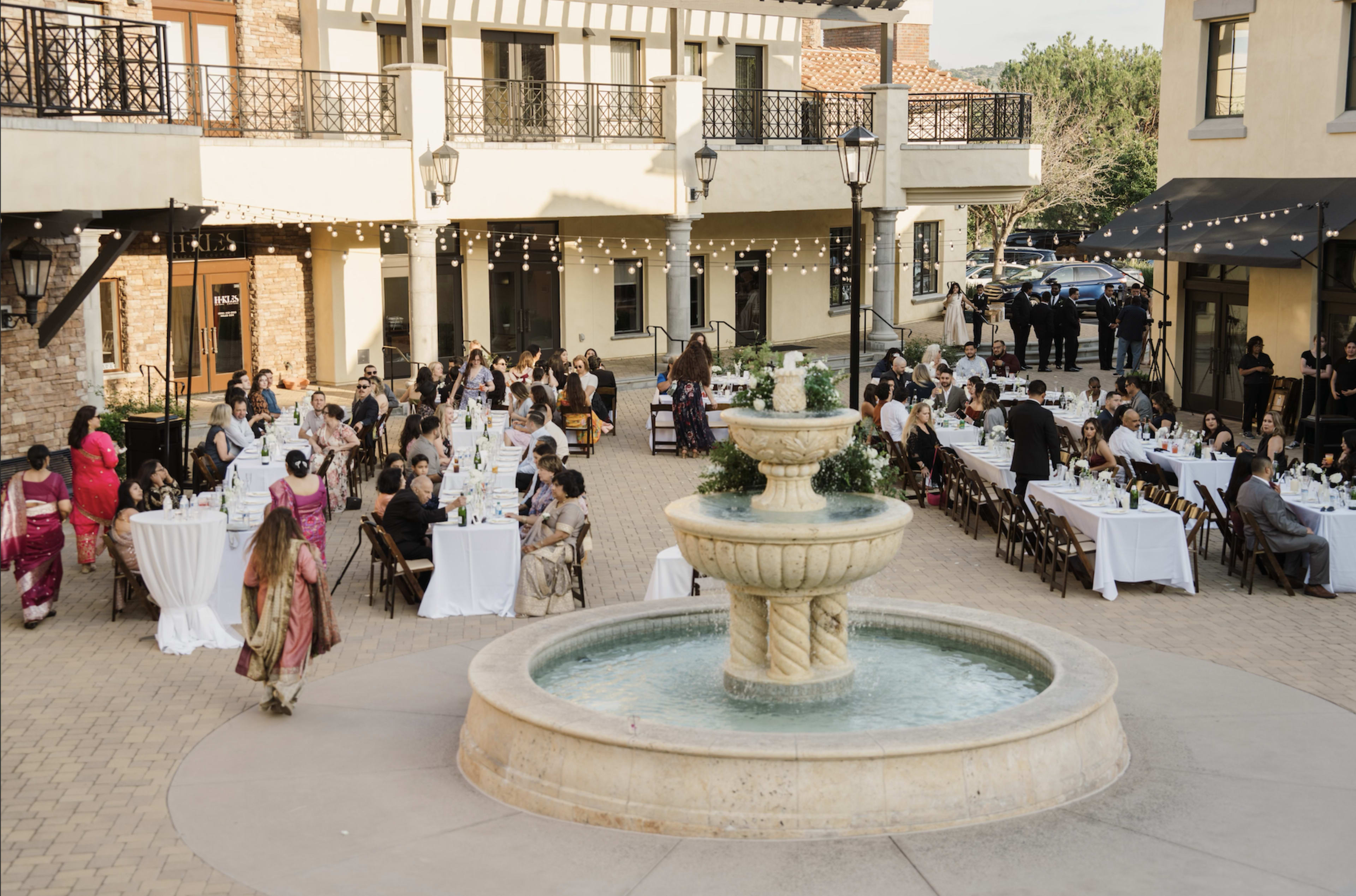 A large outdoor event is hosted in a courtyard featuring a central fountain, with guests seated at tables arranged around the area under string lights.