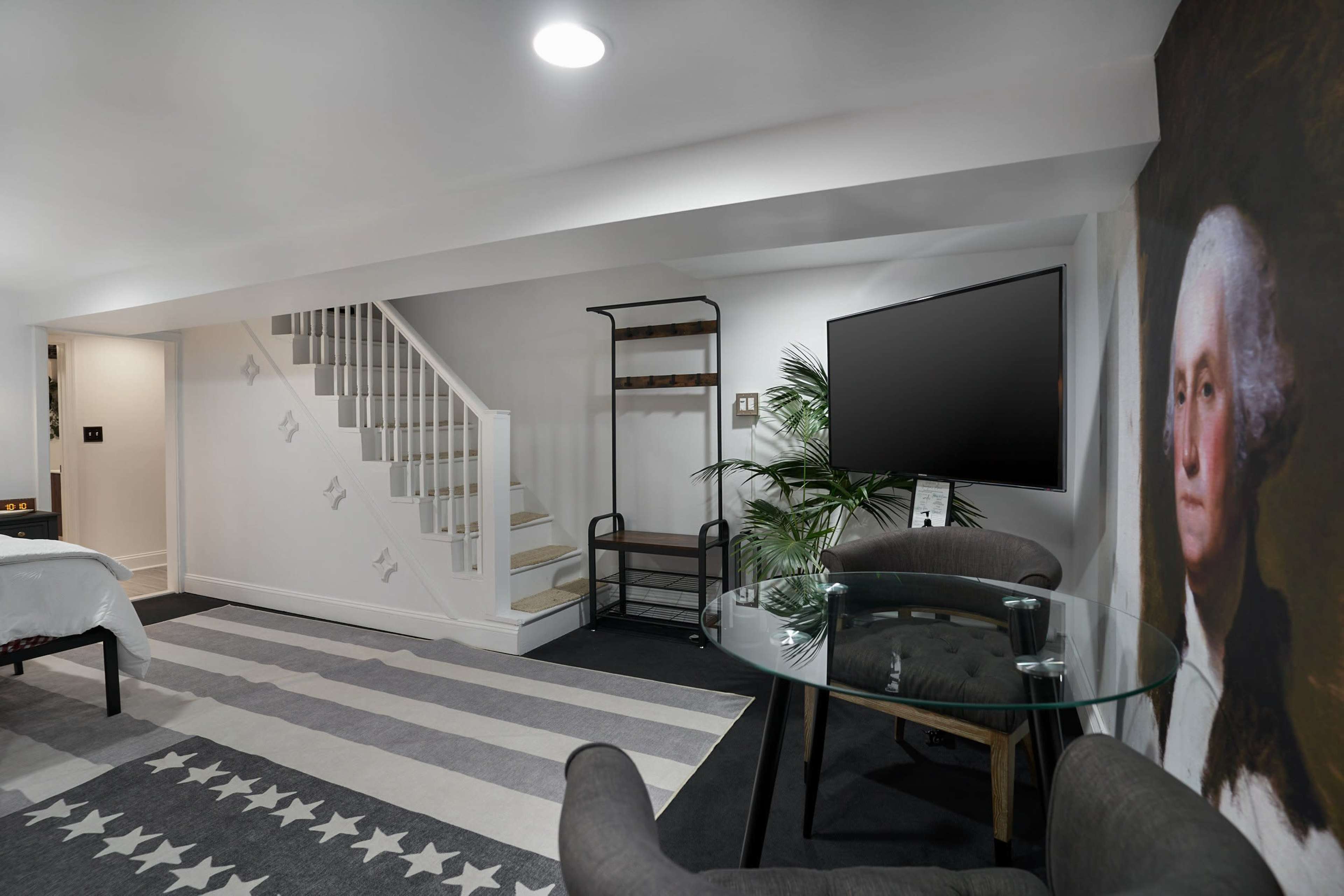 The image shows a modern basement room with a staircase, a large TV mounted on the wall, a glass table with two chairs, and an artwork of George Washington.