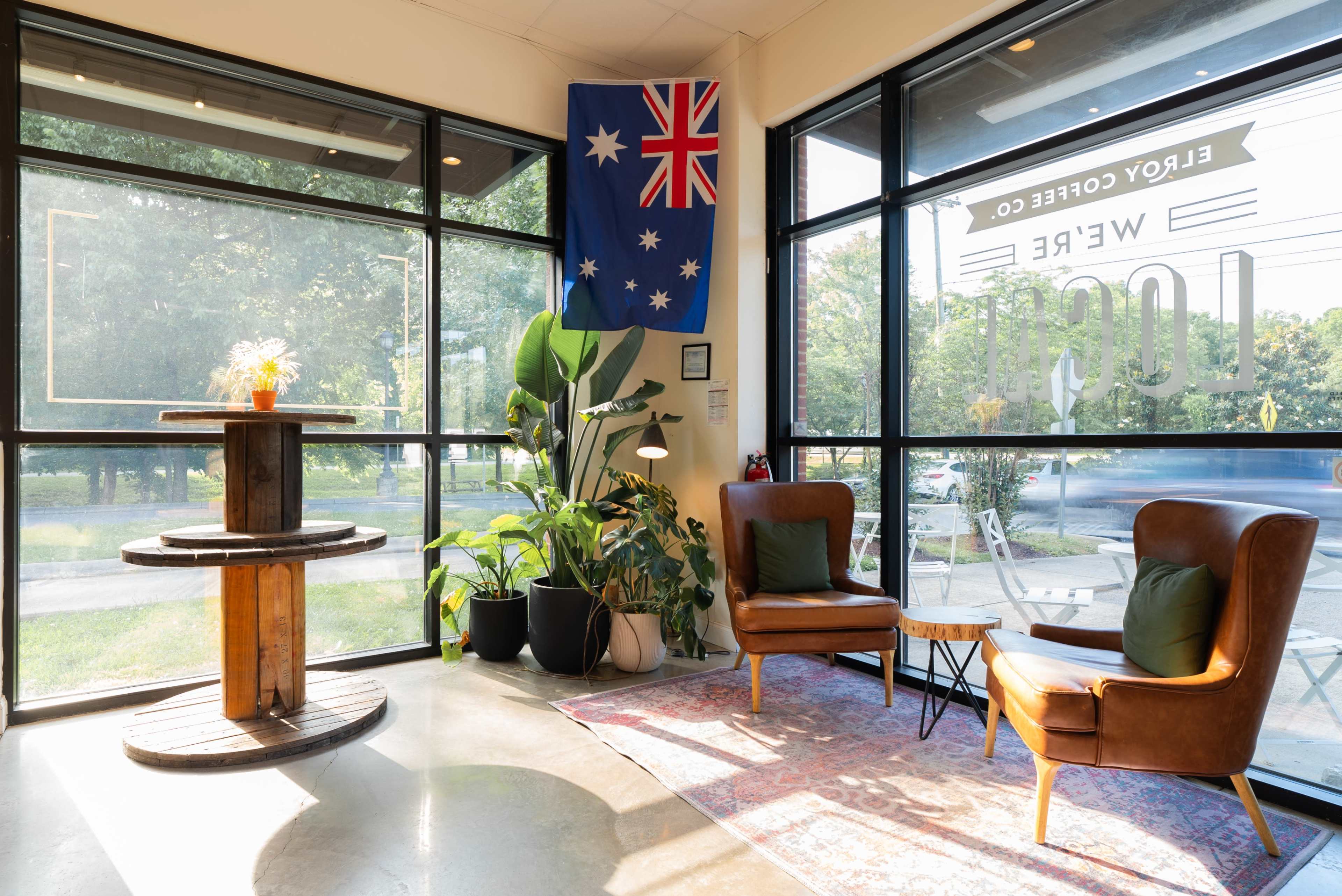 The image shows a cozy café interior with large windows, a wooden table, two brown chairs, a potted plant, and an Australian flag hanging on the wall.