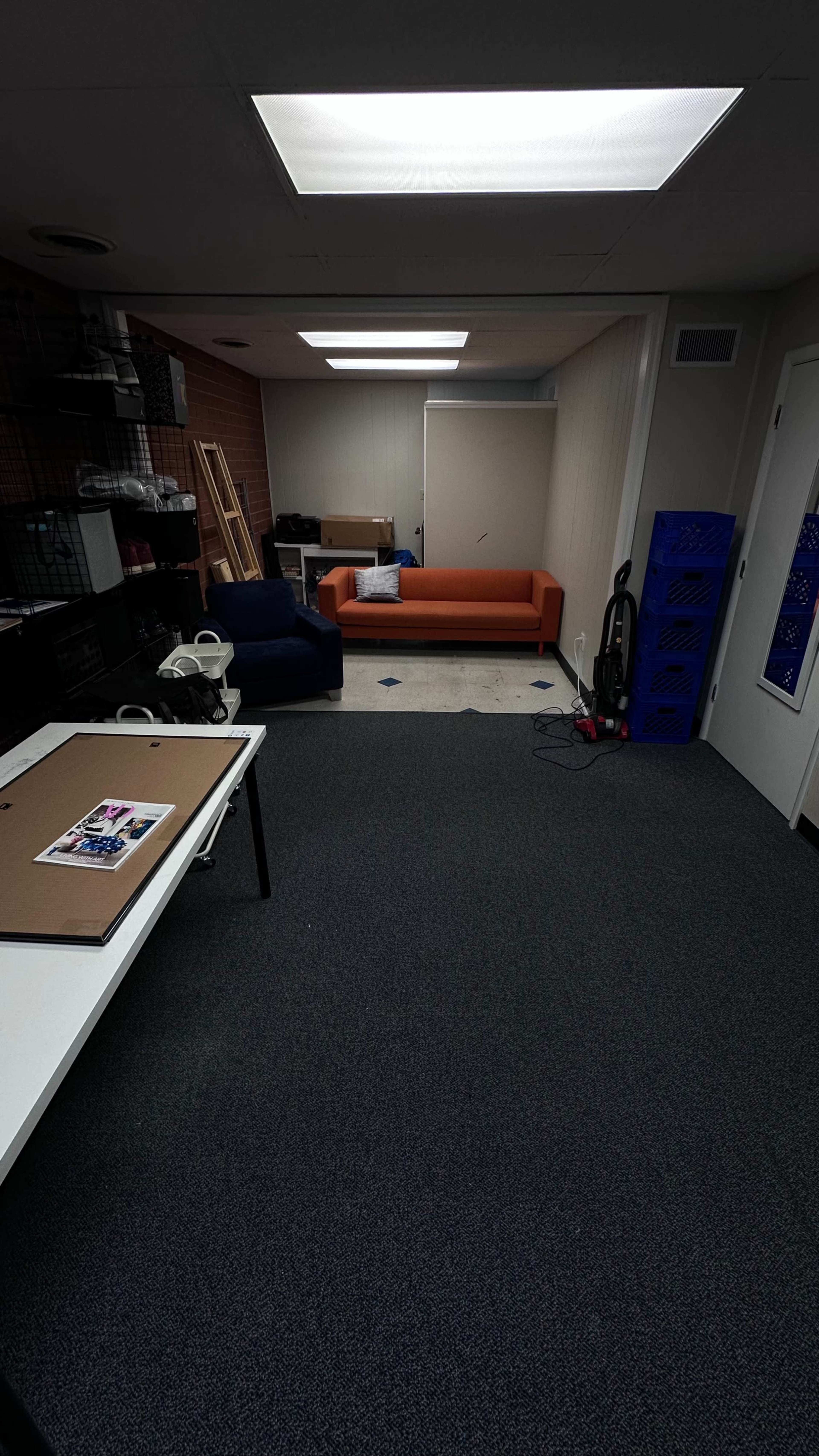 A room features an orange sofa, a blue chair, a table with art supplies, and a vacuum cleaner against the wall.