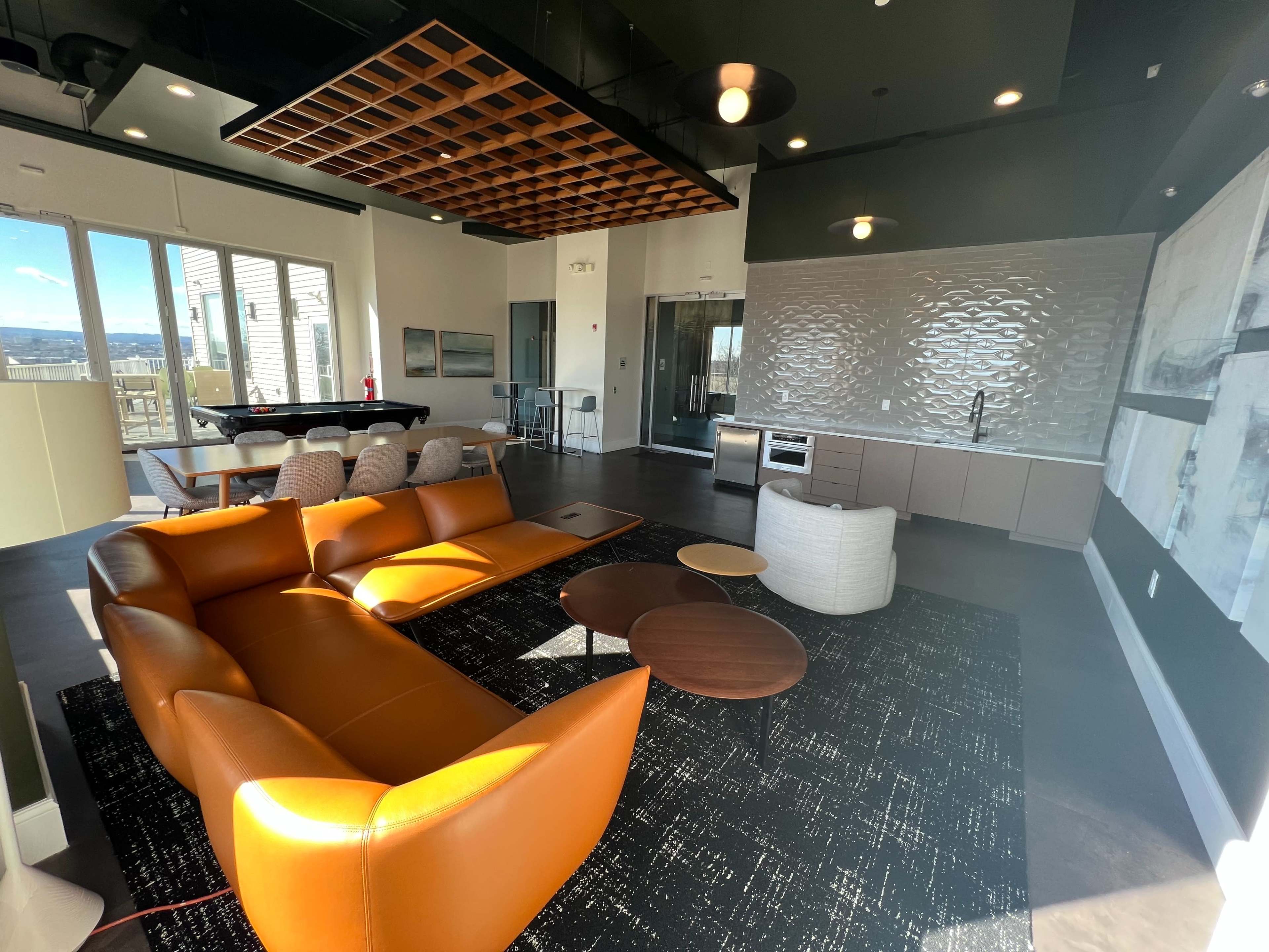 A modern living space with a brown leather sofa, round coffee tables, and a kitchenette, featuring large windows that overlook a scenic view.
