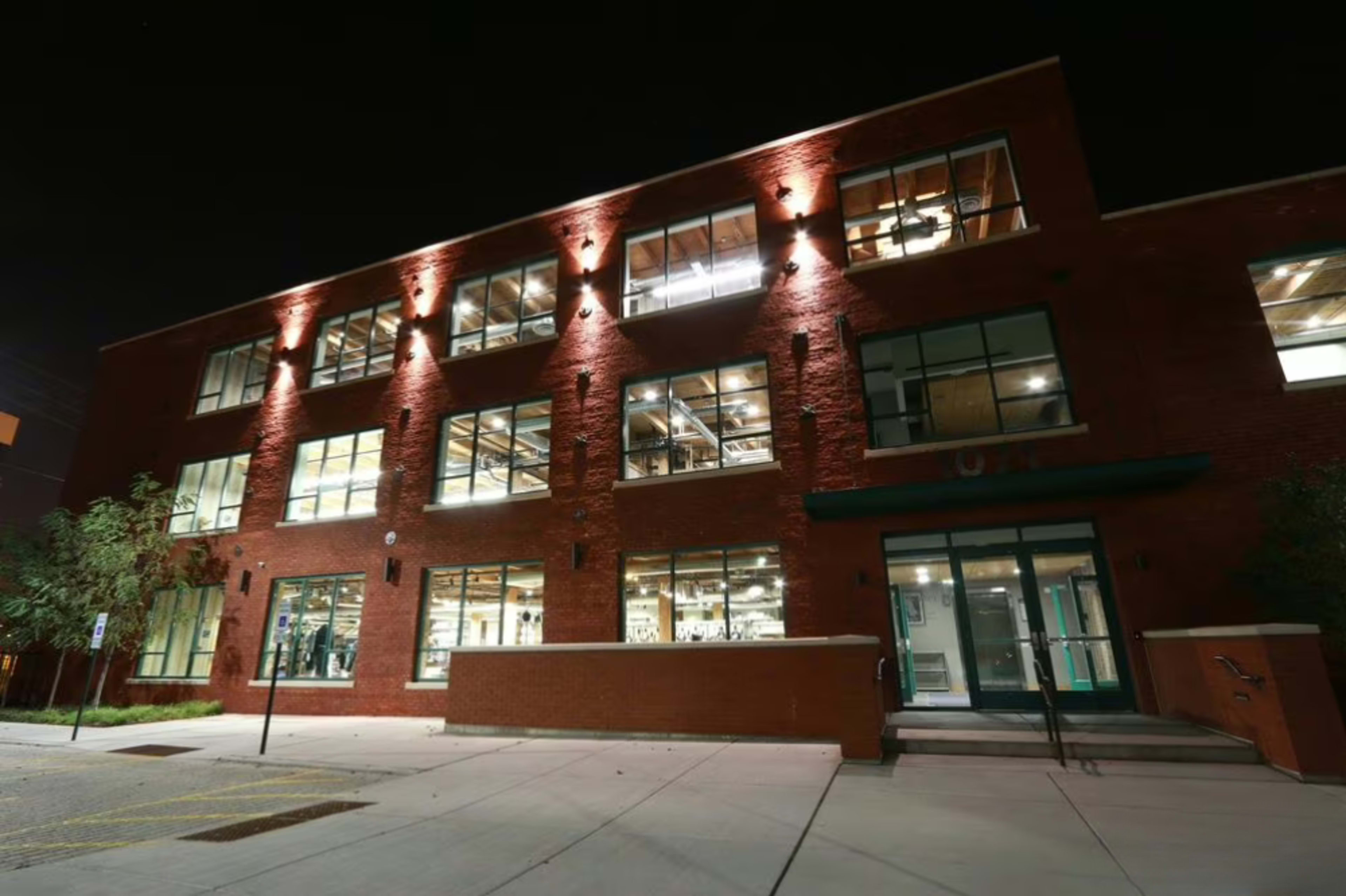 The image shows a modern brick building with large windows illuminated at night, featuring a clean entrance and surrounding landscaping.