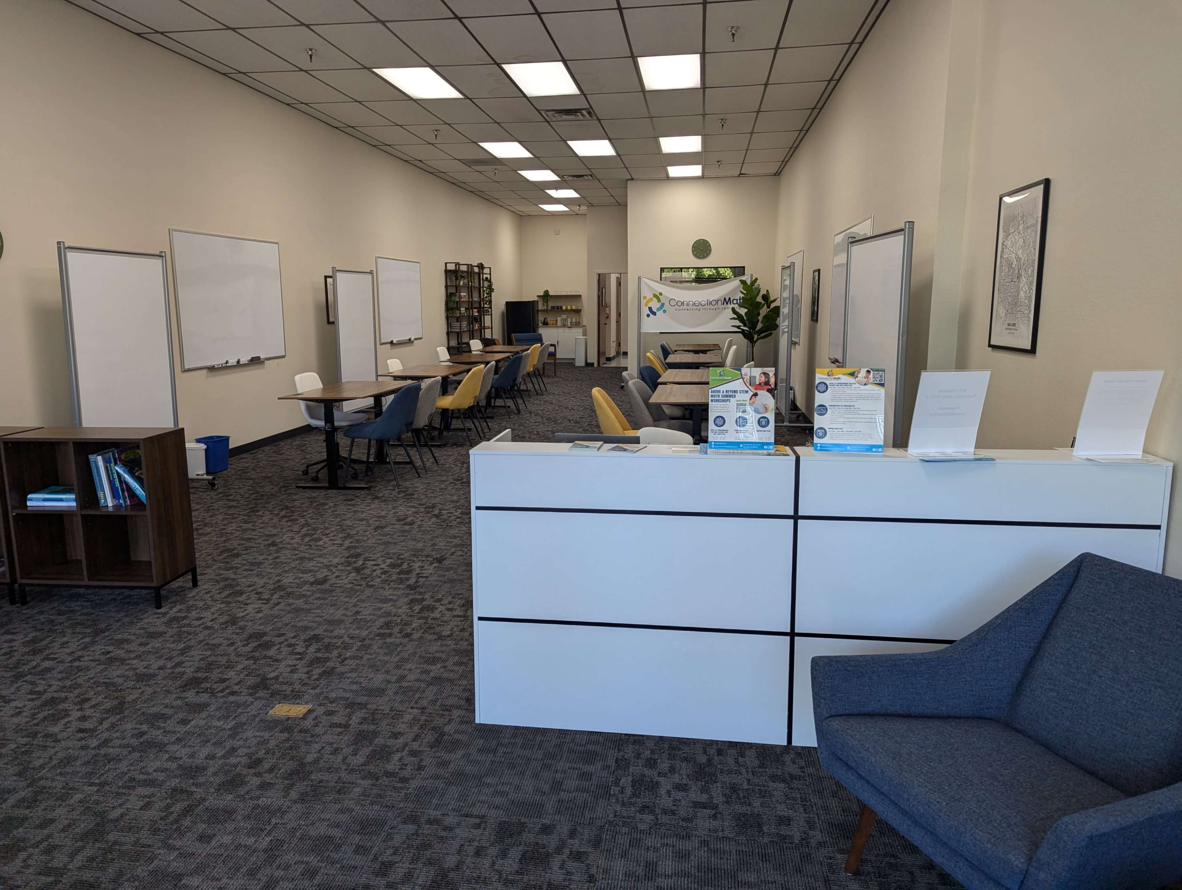 A modern office space with a reception desk in the foreground, lined with chairs and tables, and whiteboards on the walls.