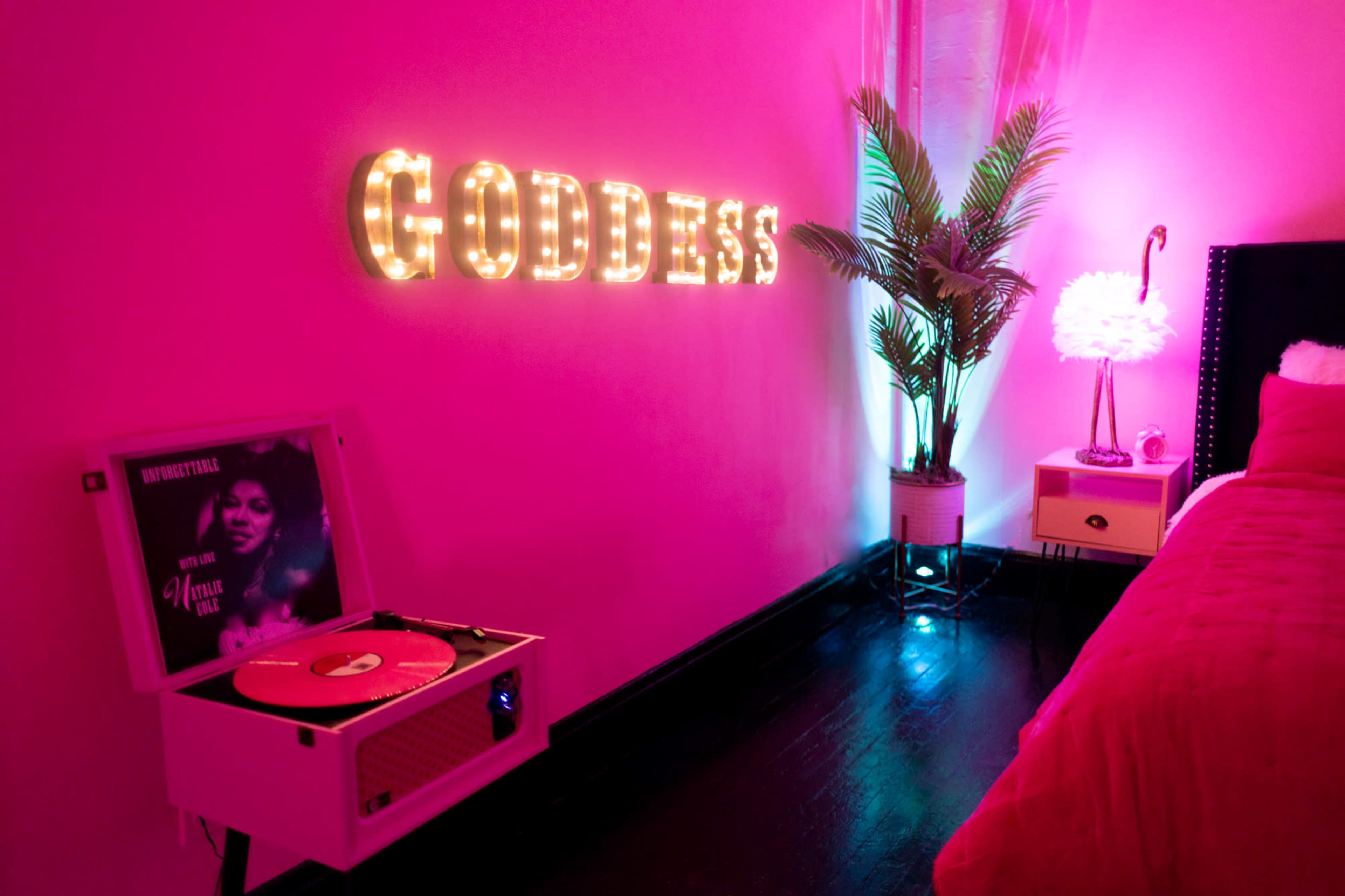 A pink-themed room features a lit "GODDESS" sign, a record player, and a decorative plant beside a bed.