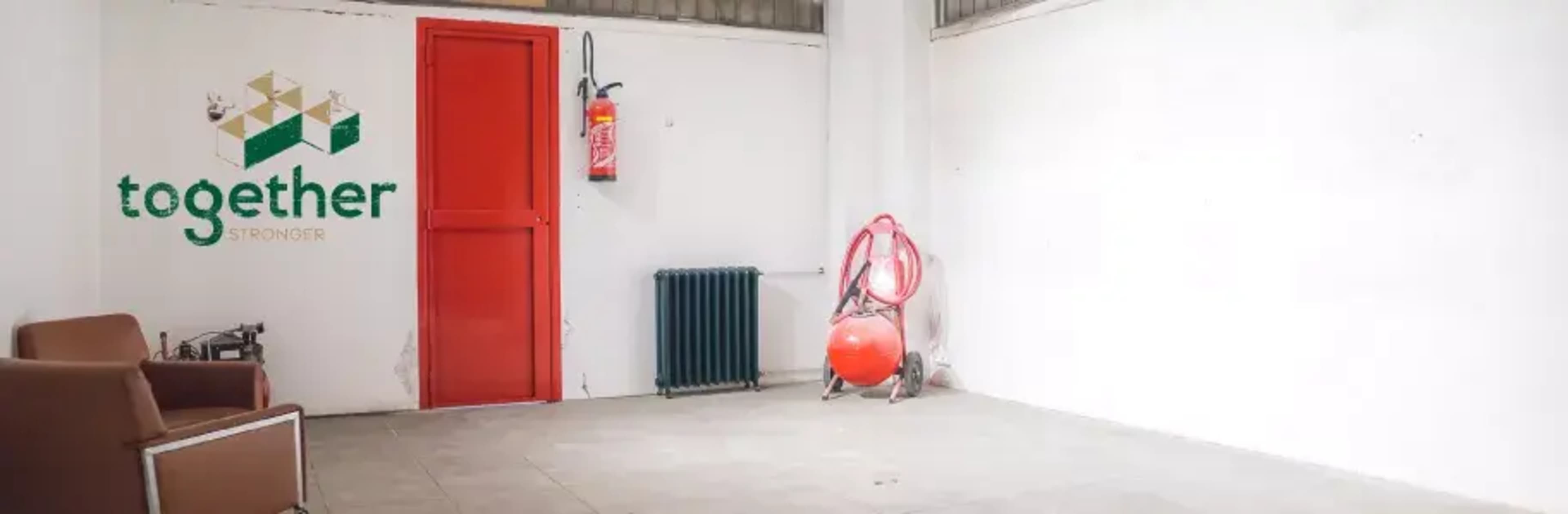 The image shows a spacious, empty room with a red door, a radiator, and a fire extinguisher mounted on the wall, along with a brown armchair and an orange object in the corner.