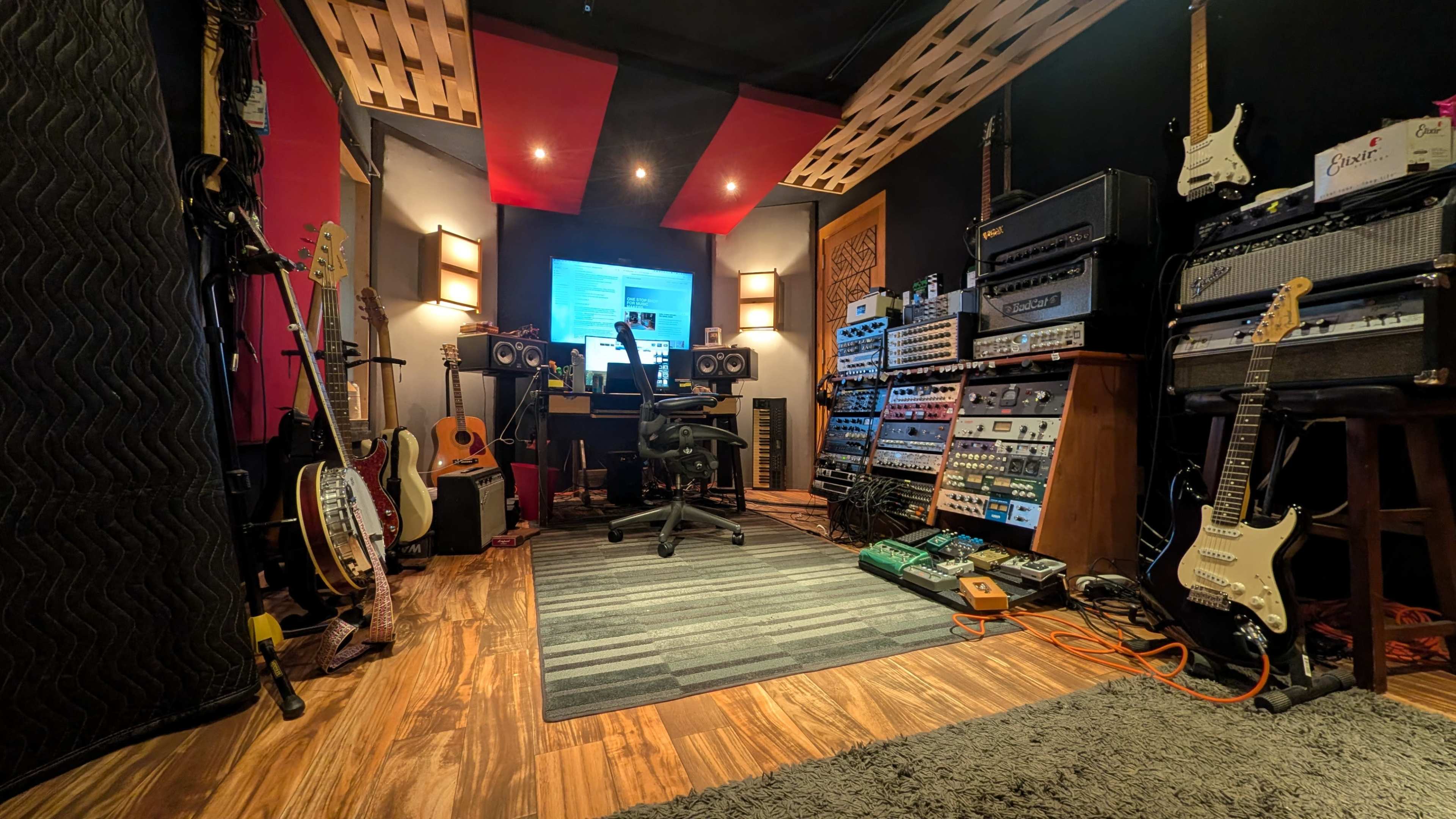 The image shows a music recording studio with various instruments, amplifiers, and sound equipment arranged around a central workstation.