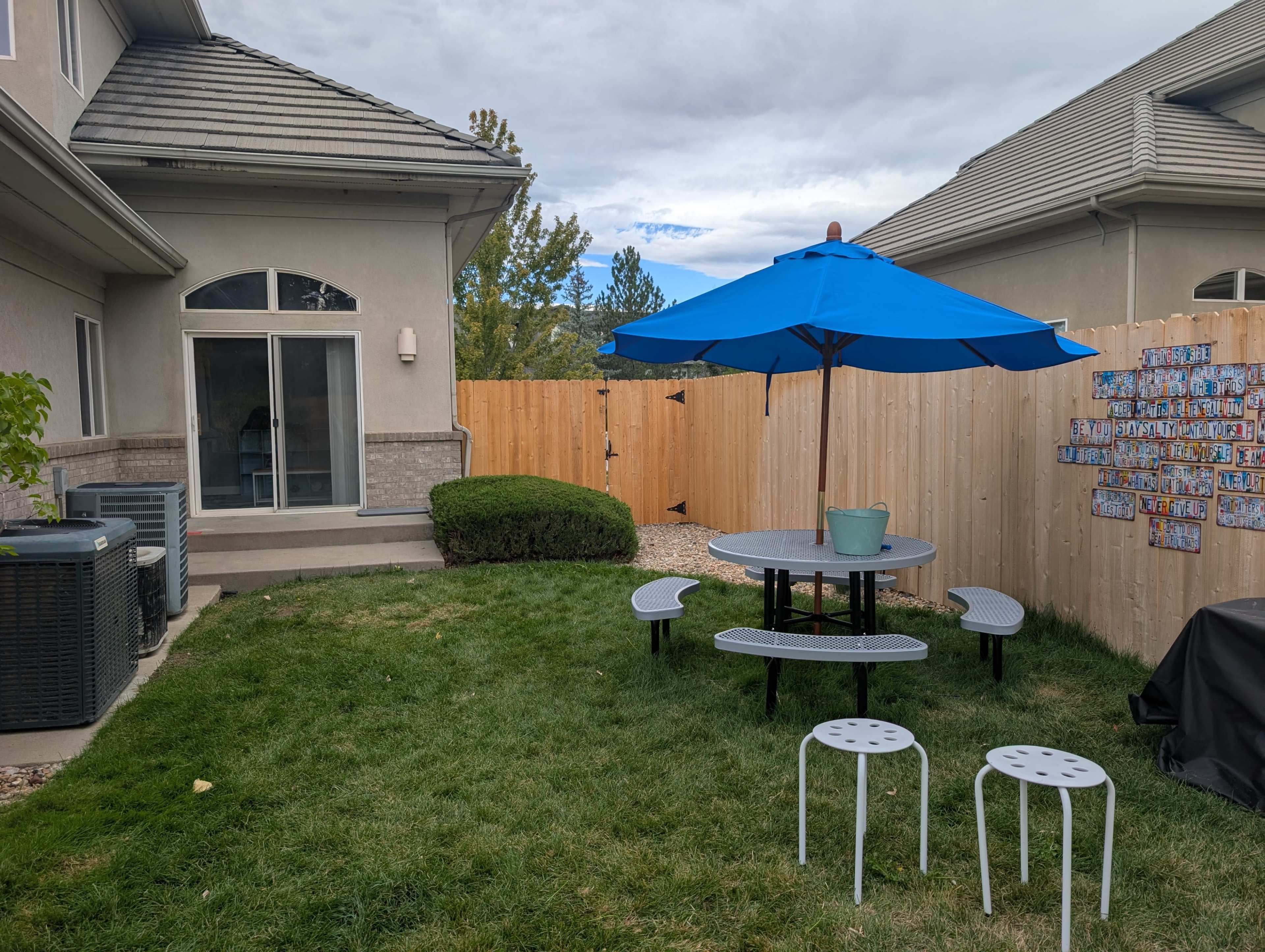 The image shows a backyard with a circular picnic table under a large blue umbrella, surrounded by green grass, a wooden privacy fence, and a house on one side.