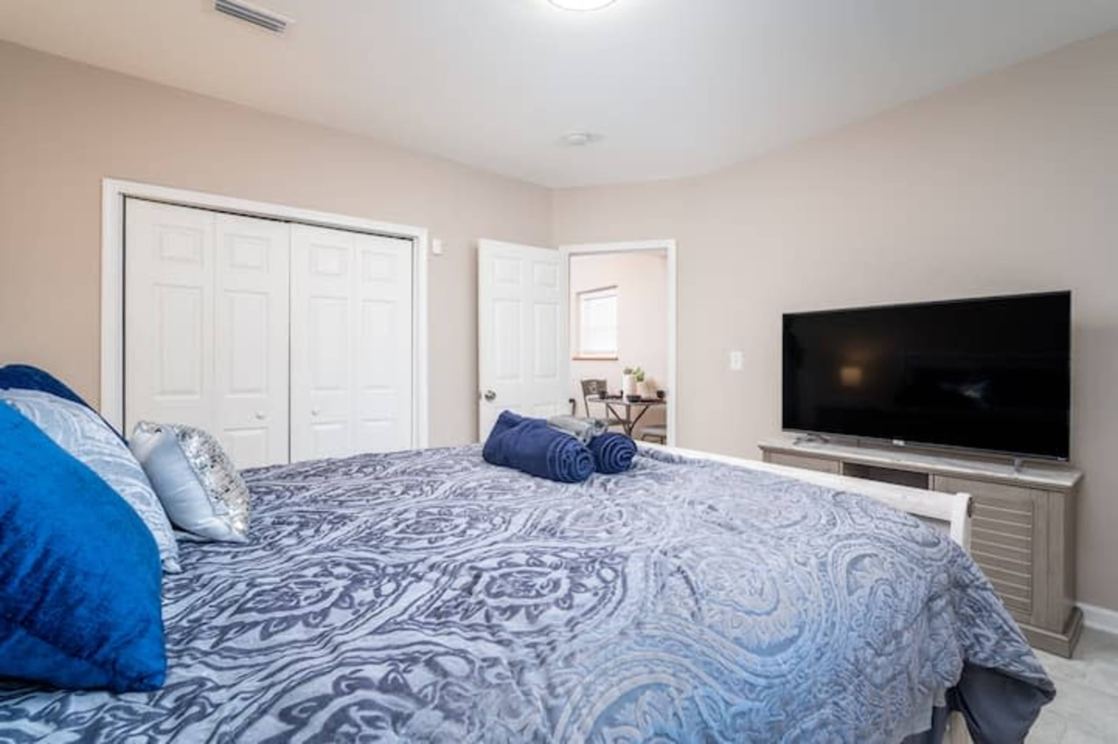 A spacious bedroom features a large bed with a patterned comforter, neatly rolled towels, a television on a stand, and a doorway leading to another room.