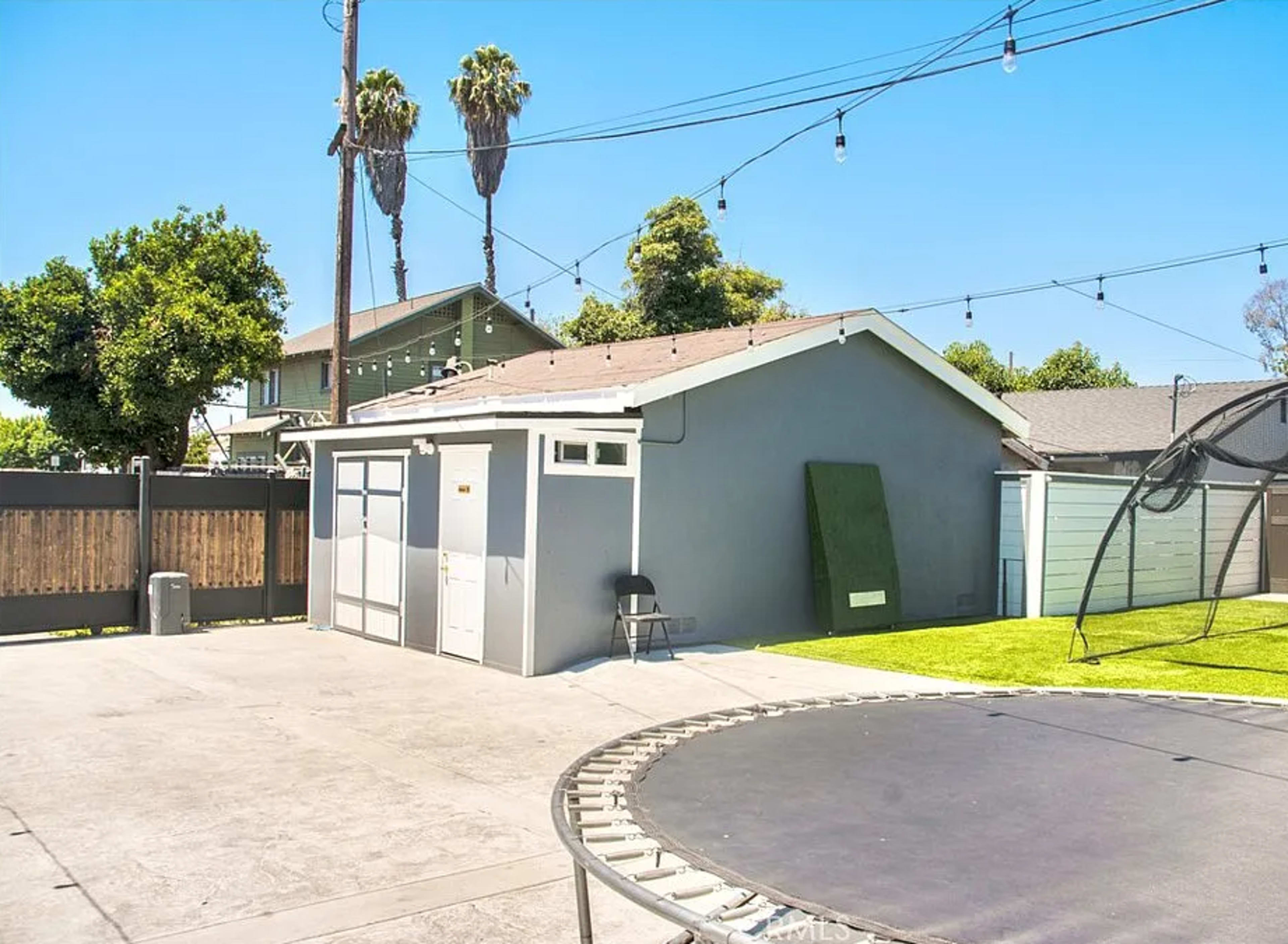 A gray shed is positioned beside a trampoline in a backyard, with palm trees and a two-story house visible in the background.