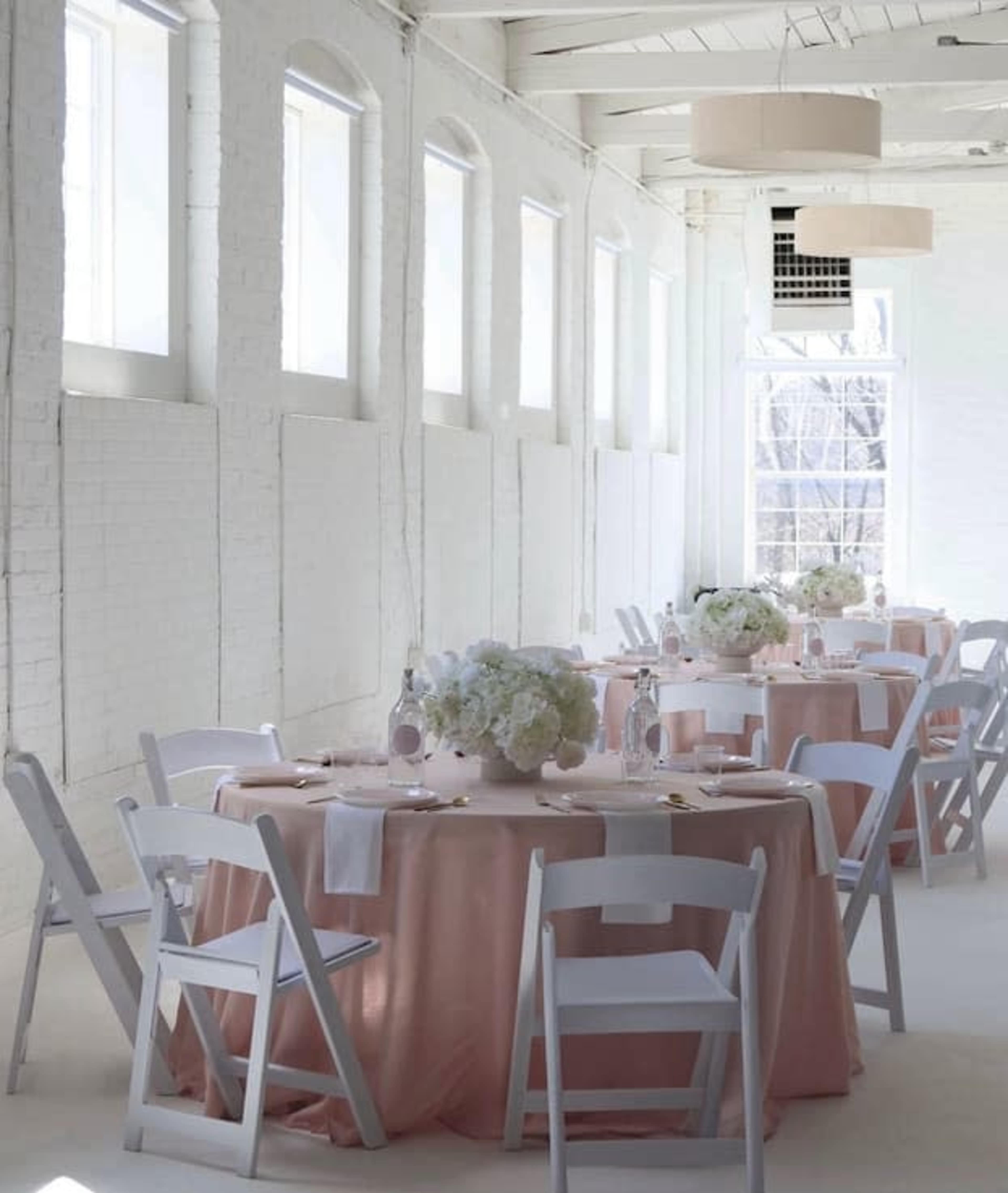 The image shows a well-lit event space with several round tables covered in pale pink tablecloths, each adorned with floral centerpieces, surrounded by white folding chairs.