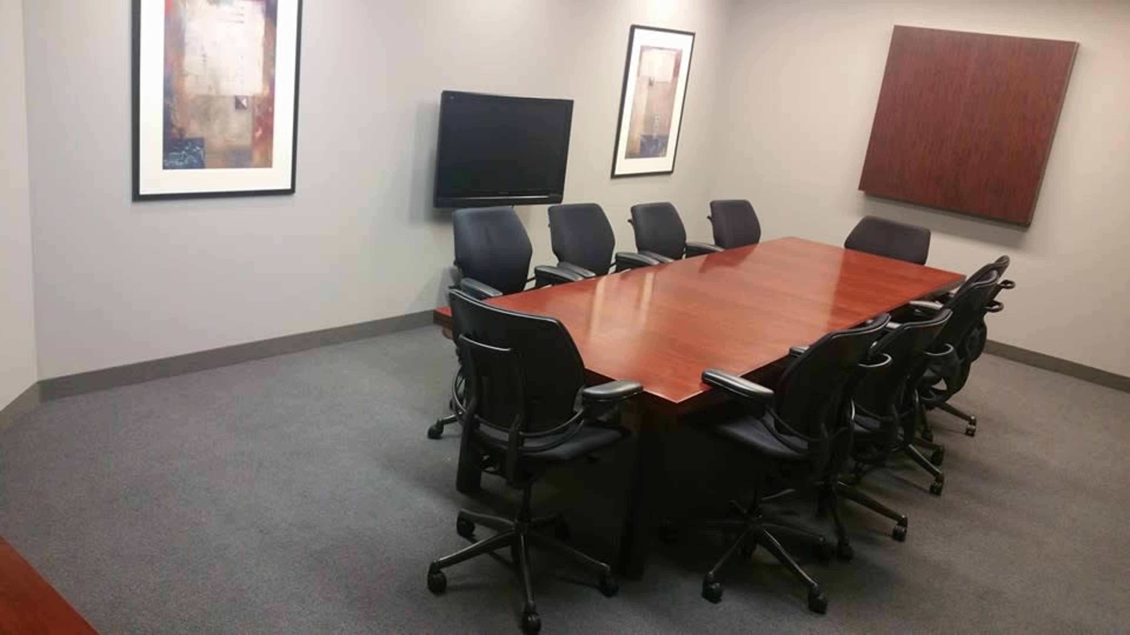 A conference room features a large wooden table surrounded by black rolling chairs, with two framed artworks and a television mounted on the wall.