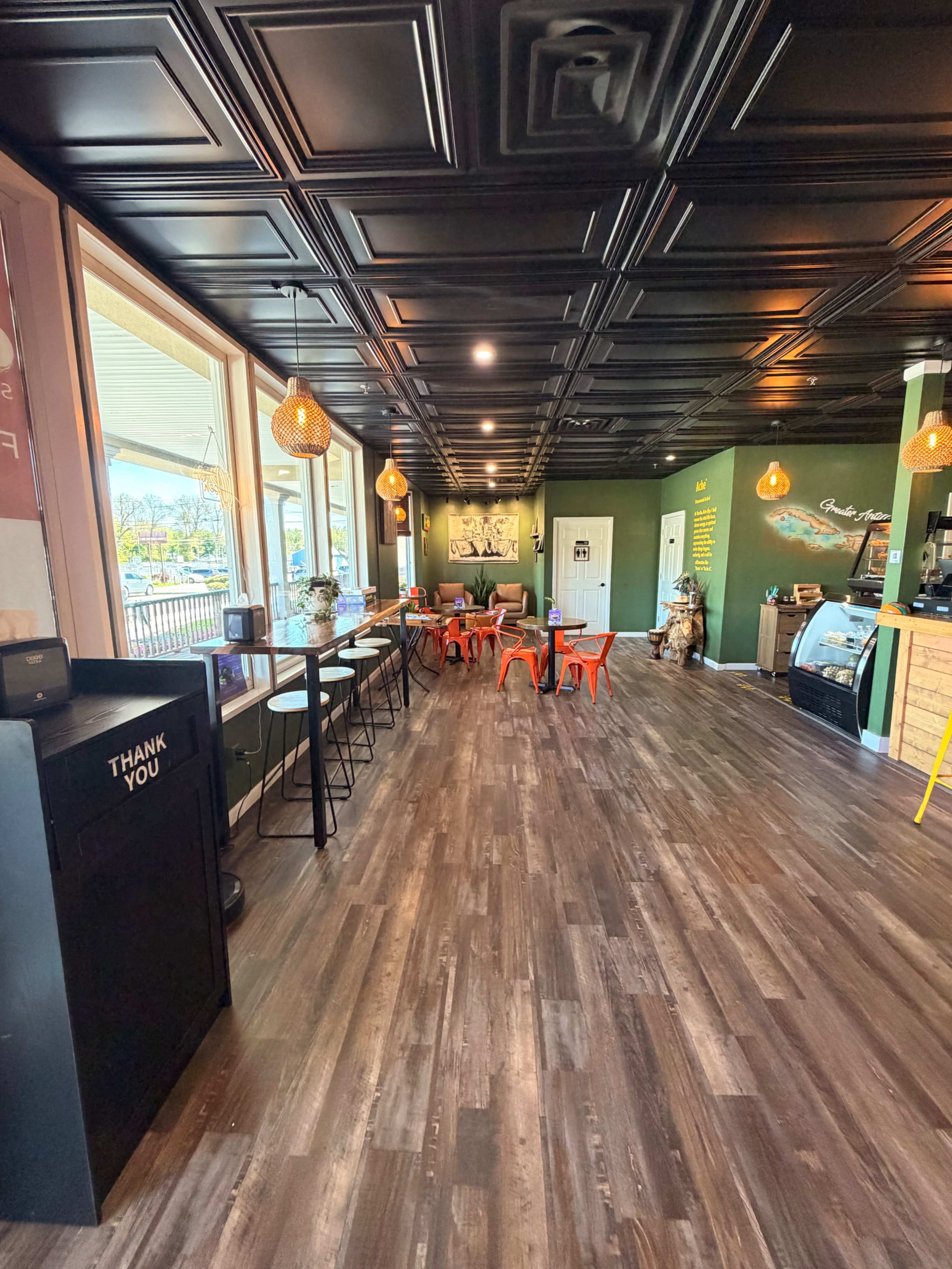The image shows a modern café interior with a wooden floor, green walls, high tables, and bright orange chairs, illuminated by decorative pendant lighting.