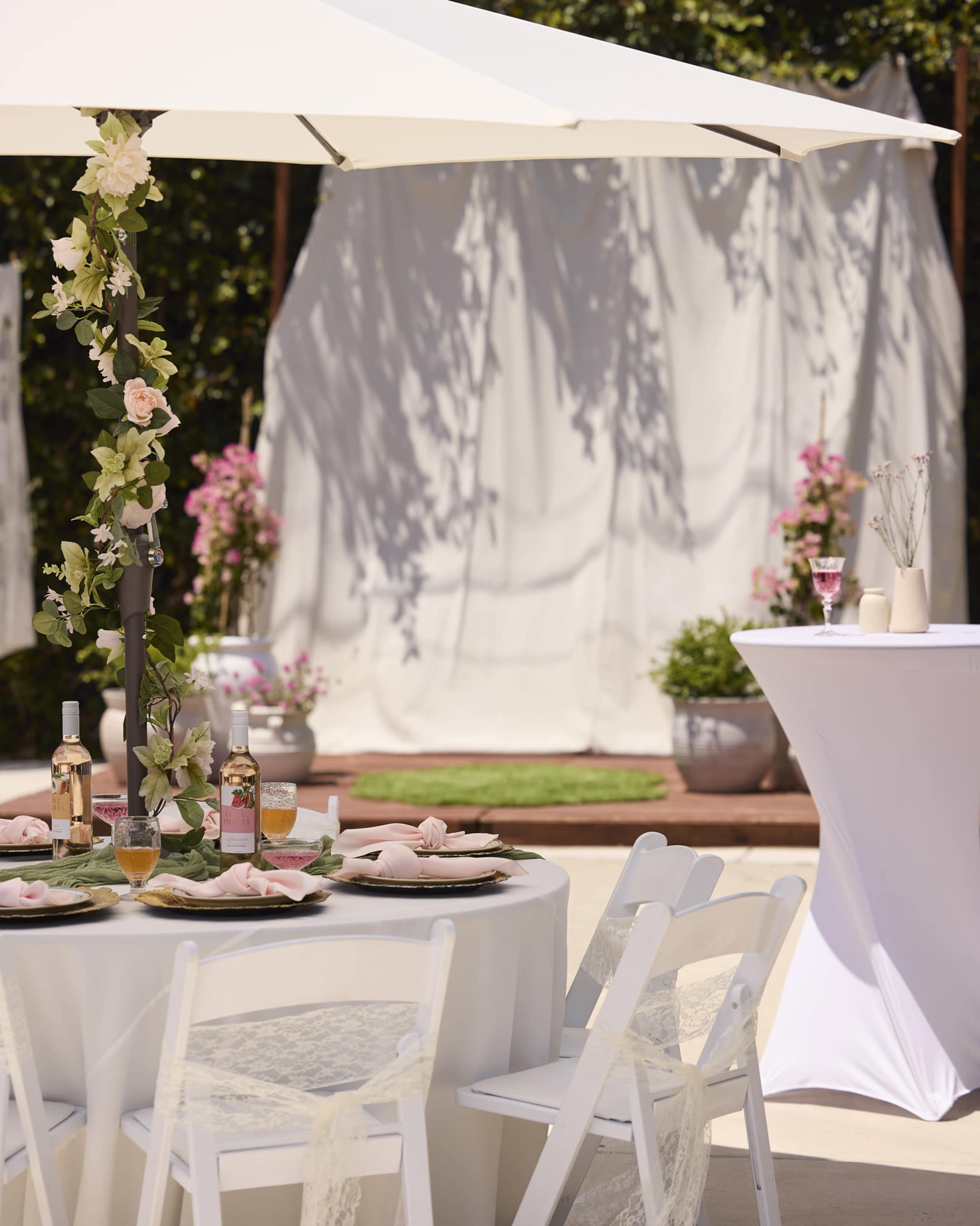A round table with white tablecloths is set for a meal, featuring drinks and decorative flowers under a white umbrella, with a backdrop of draped fabric and potted plants.