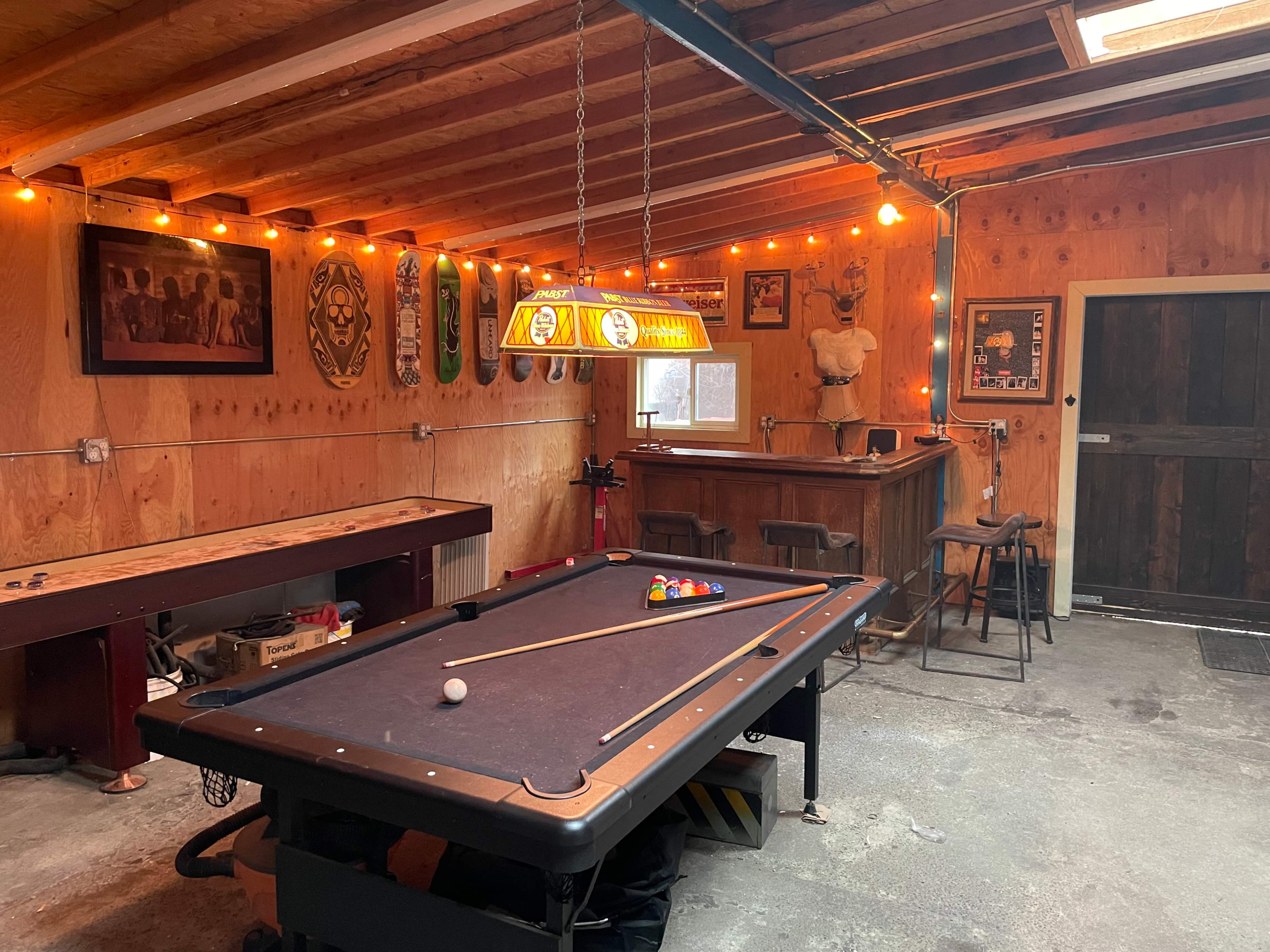 A rustic game room with a pool table, bar area, and wooden walls illuminated by string lights.