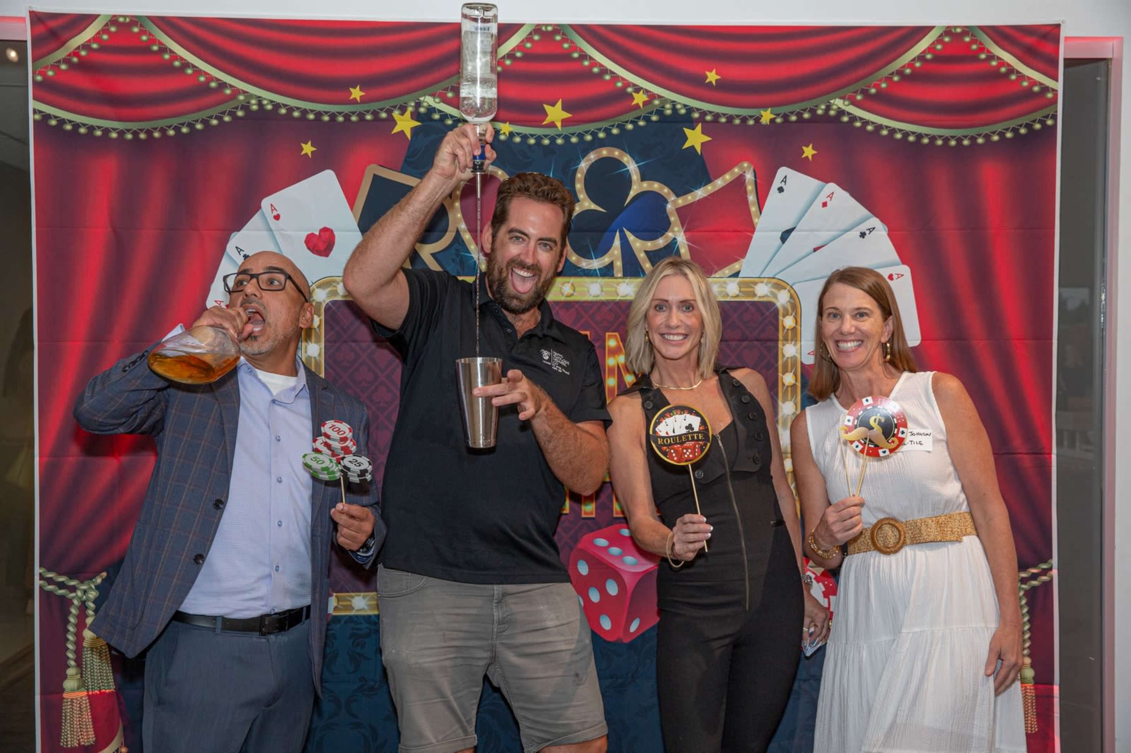 A group of four people poses with drinks and props in front of a red and gold casino-themed backdrop.