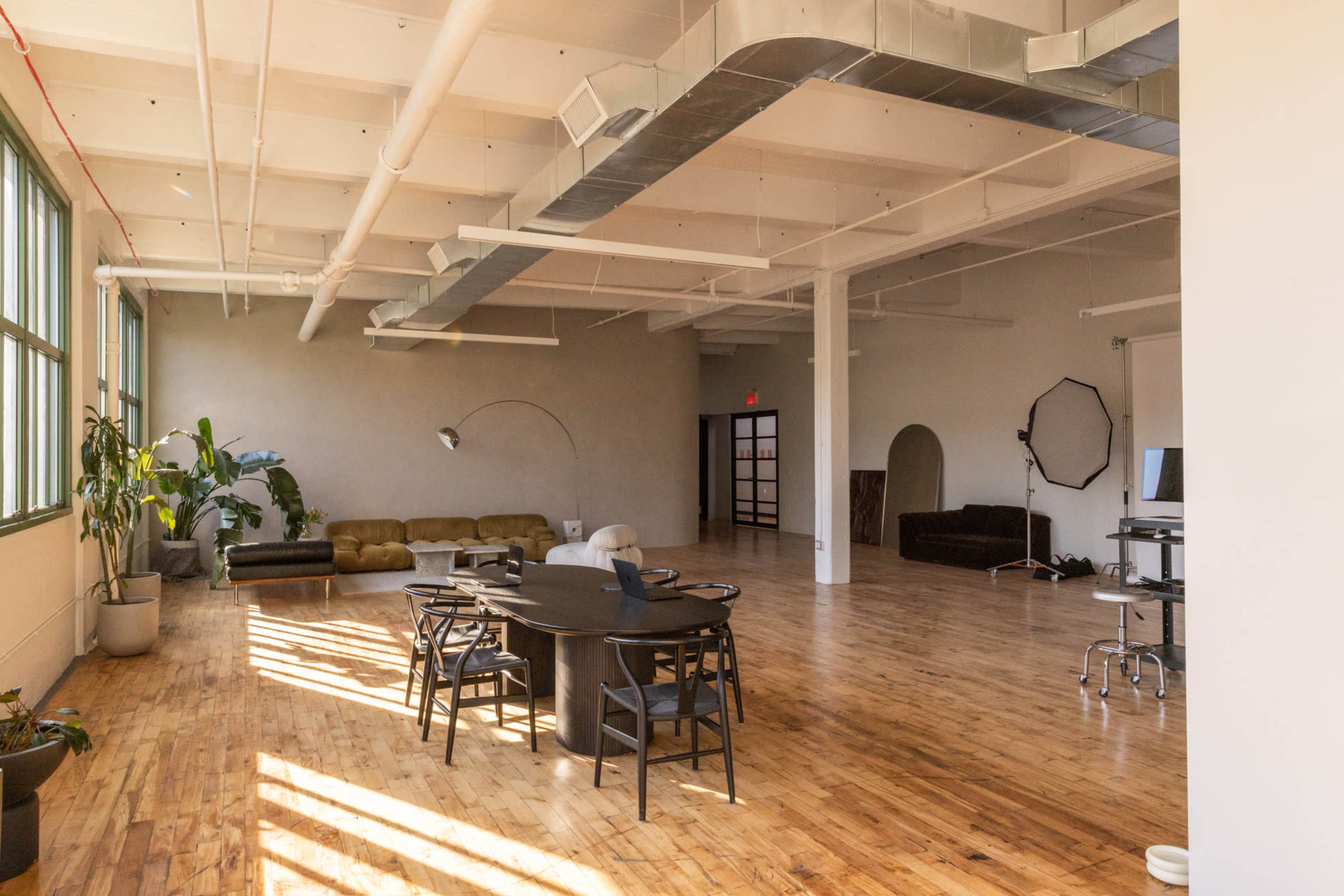 The image shows a spacious, well-lit interior with hardwood flooring, a large round table set for meetings, a couch along one wall, and various plants in decorative pots.