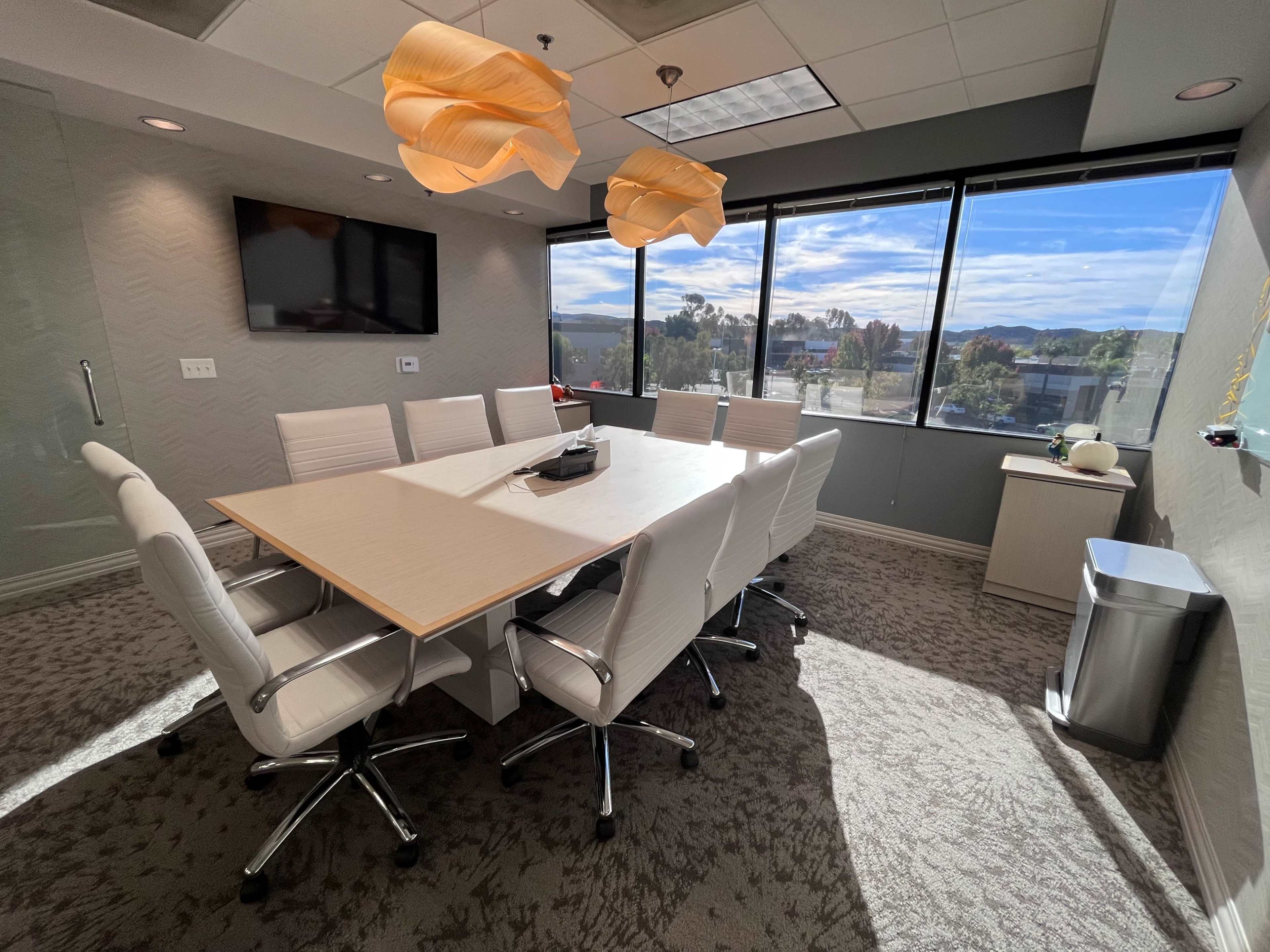 A modern conference room features a large table surrounded by white chairs, with windows offering a view of the outdoors and contemporary lighting fixtures above.
