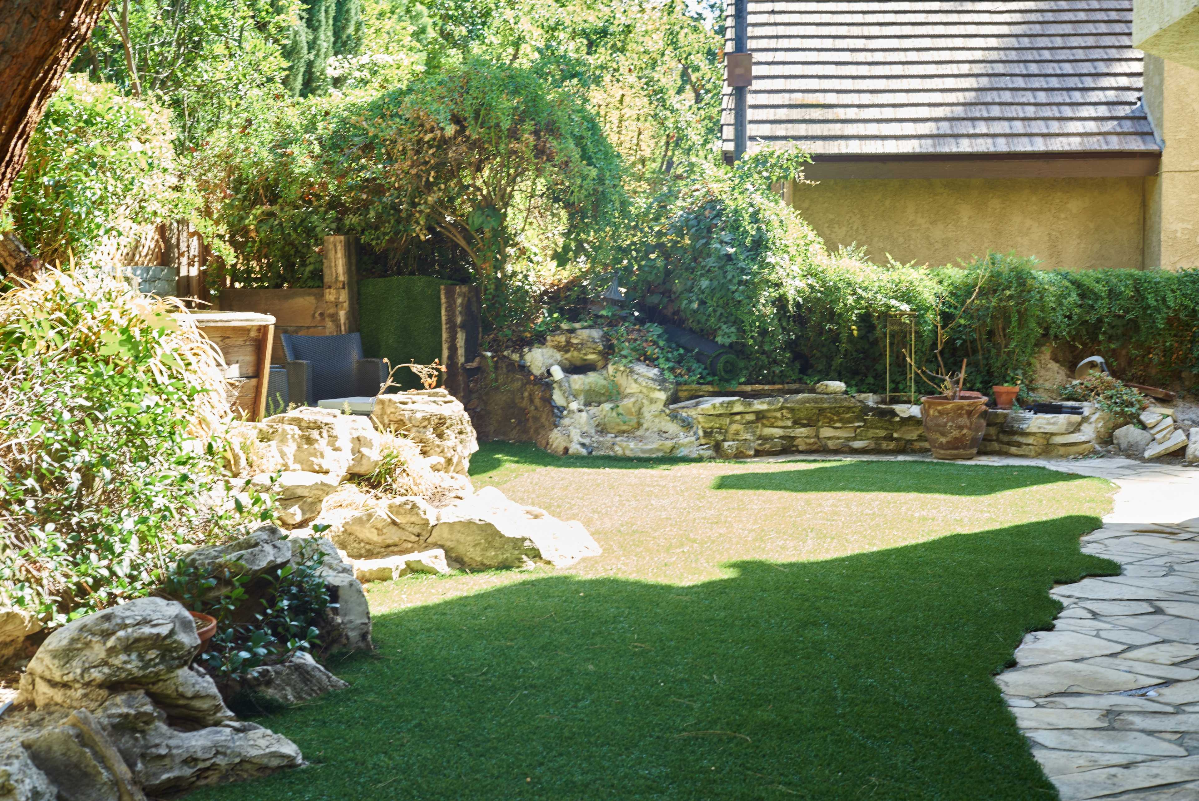 The image shows a landscaped backyard featuring a grassy area surrounded by rocks, a small water feature, and a stone pathway next to a house.
