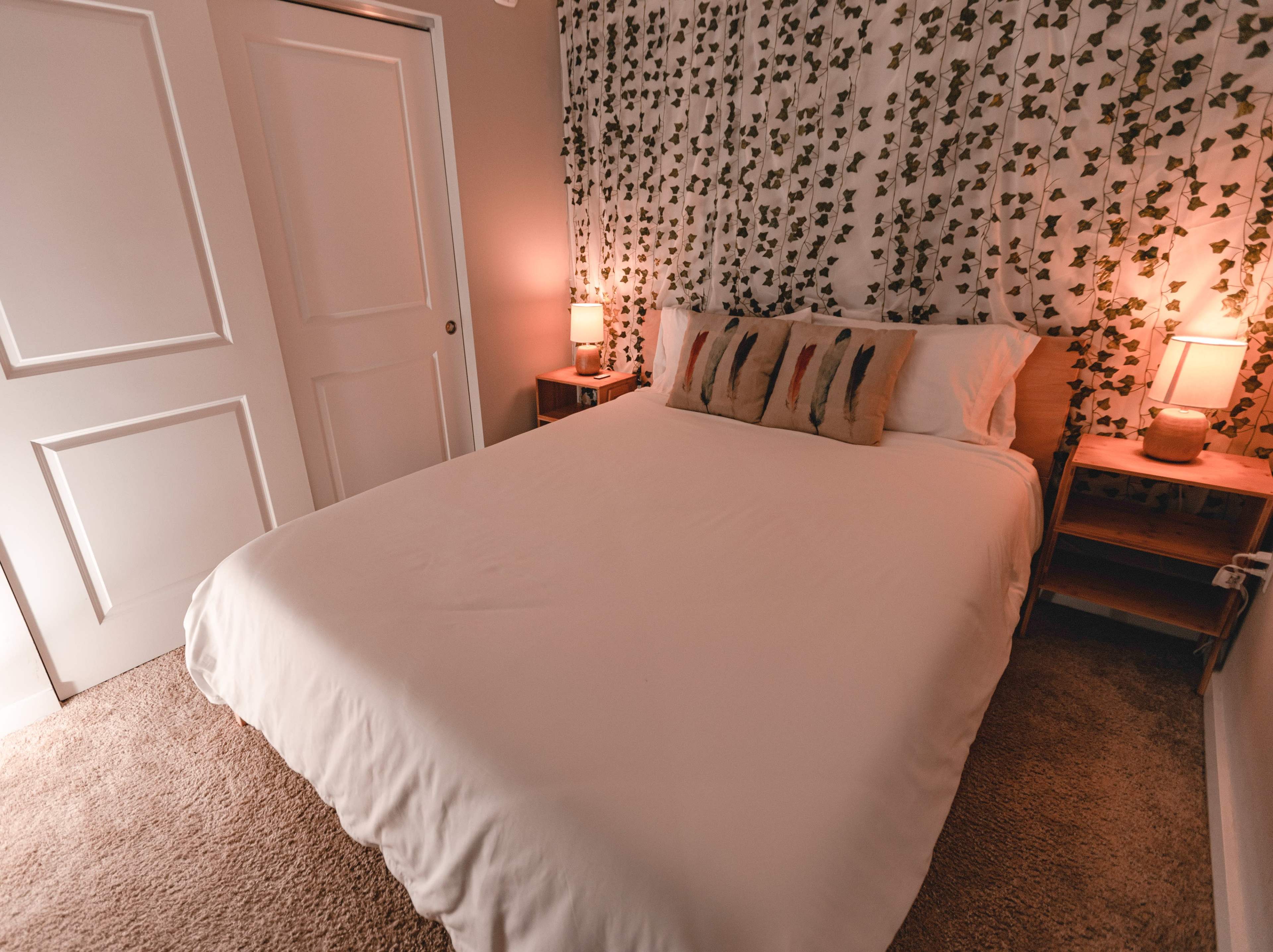 A neatly made bed in a softly lit room, with a decorative plant-patterned wall and side tables featuring lamps on either side.