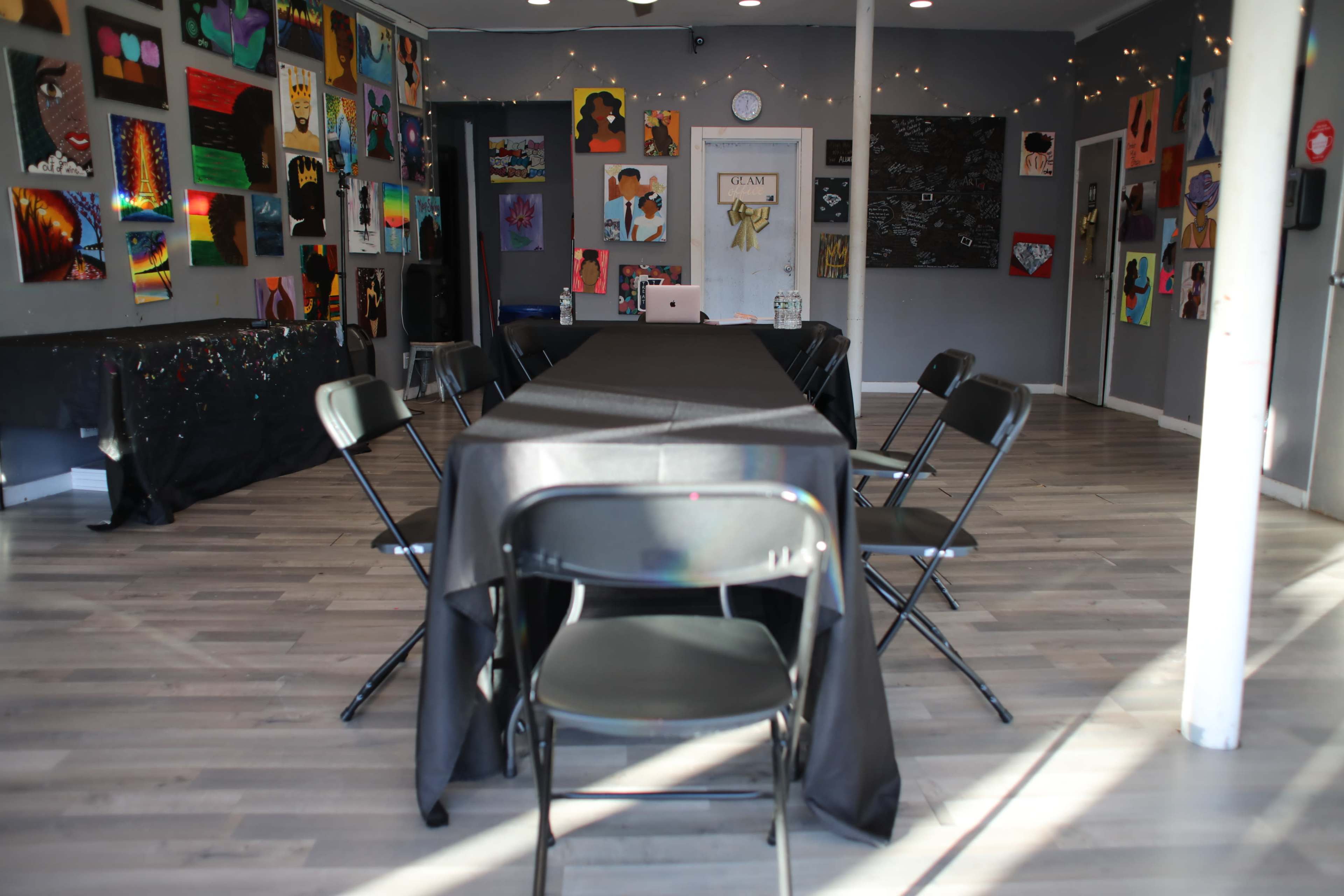 A spacious room with a long table covered in a black cloth, surrounded by folding chairs, and decorated with various colorful artworks on the walls.