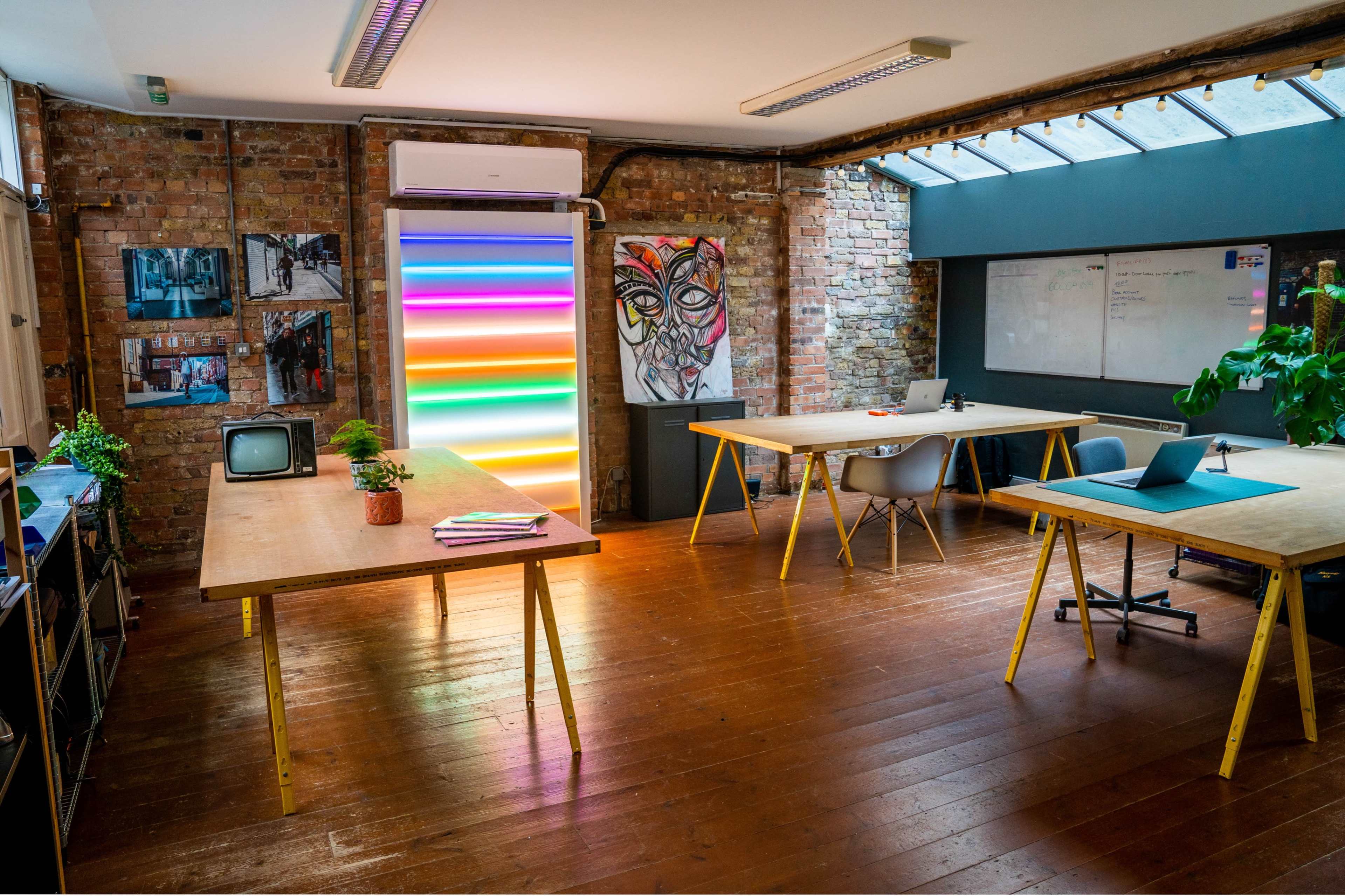 A modern workspace with wooden floors, multiple tables, a colorful neon light installation, and exposed brick walls adorned with artwork and photographs.