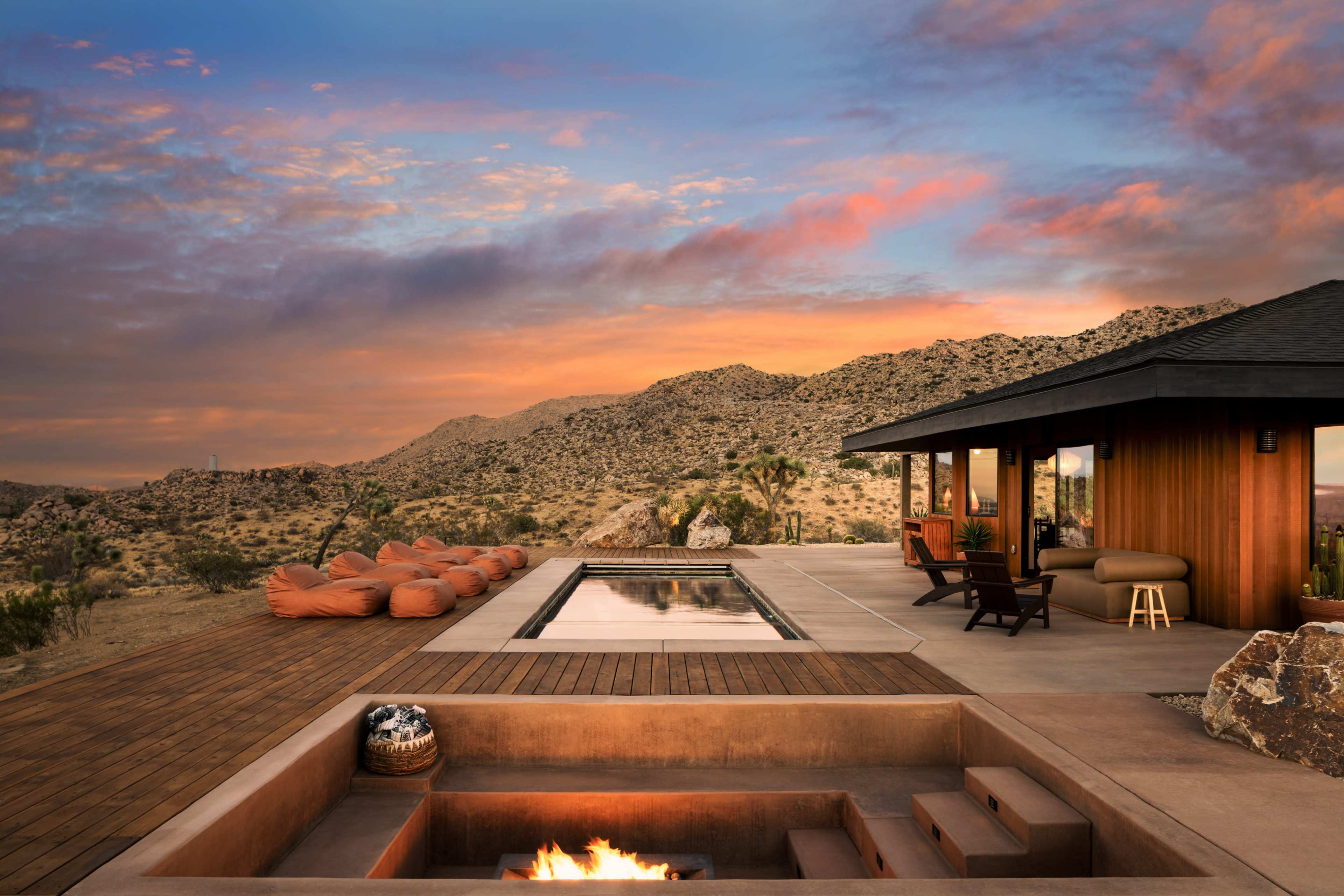 A modern home features a pool and fire pit, surrounded by desert landscapes and mountains under a colorful sunset sky.