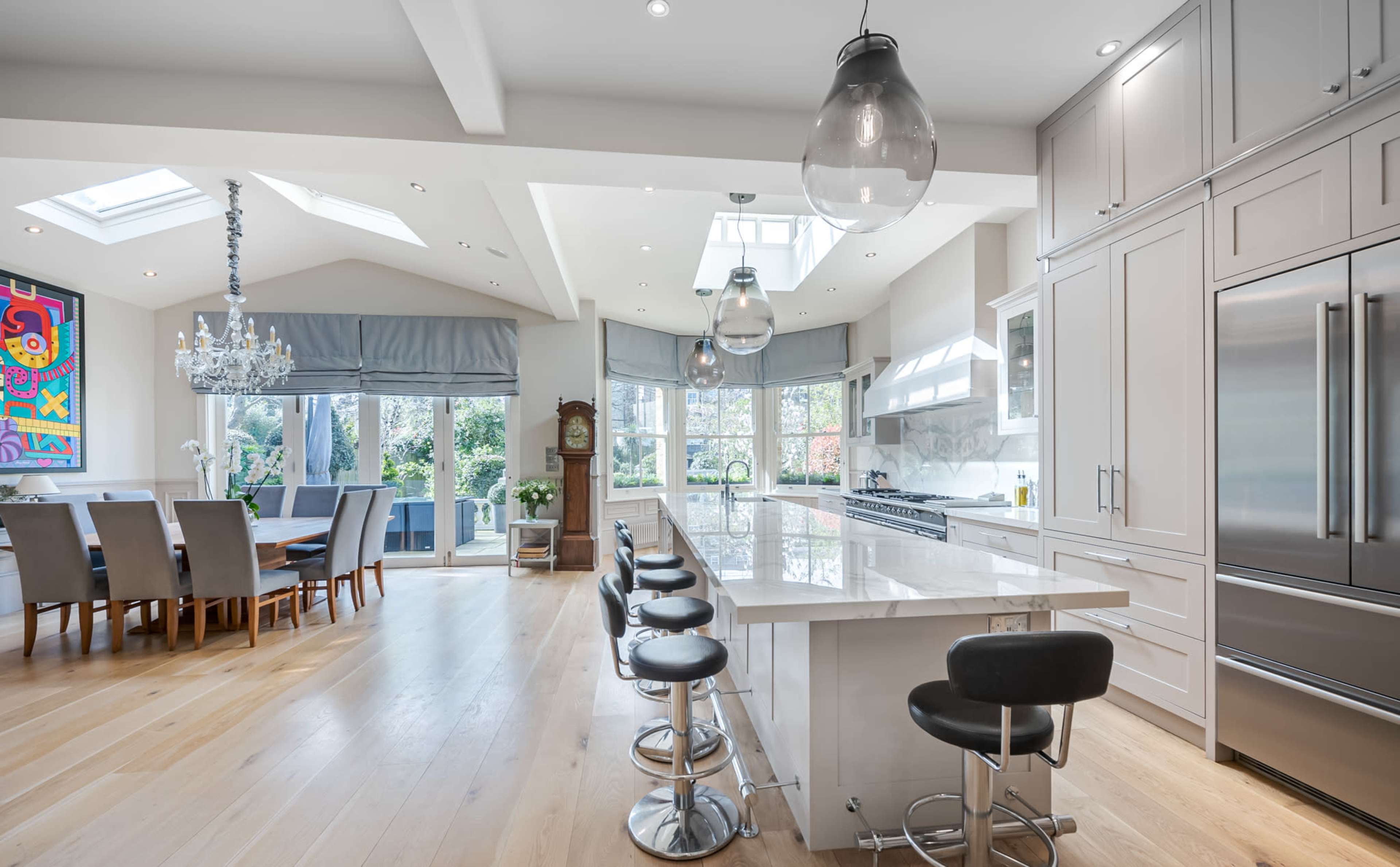 A modern kitchen features a large island with bar seating, stainless steel appliances, and a dining area with a large table, all illuminated by natural light from skylights and large windows.