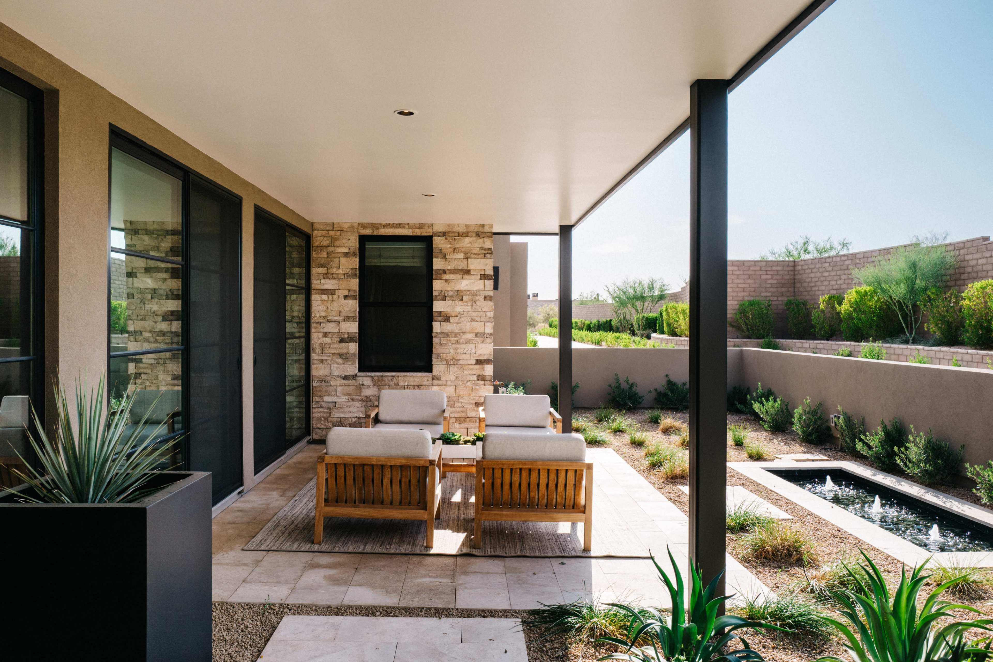 A covered patio features two wooden chairs and a small table, with a stone wall and landscaped greenery in the background.