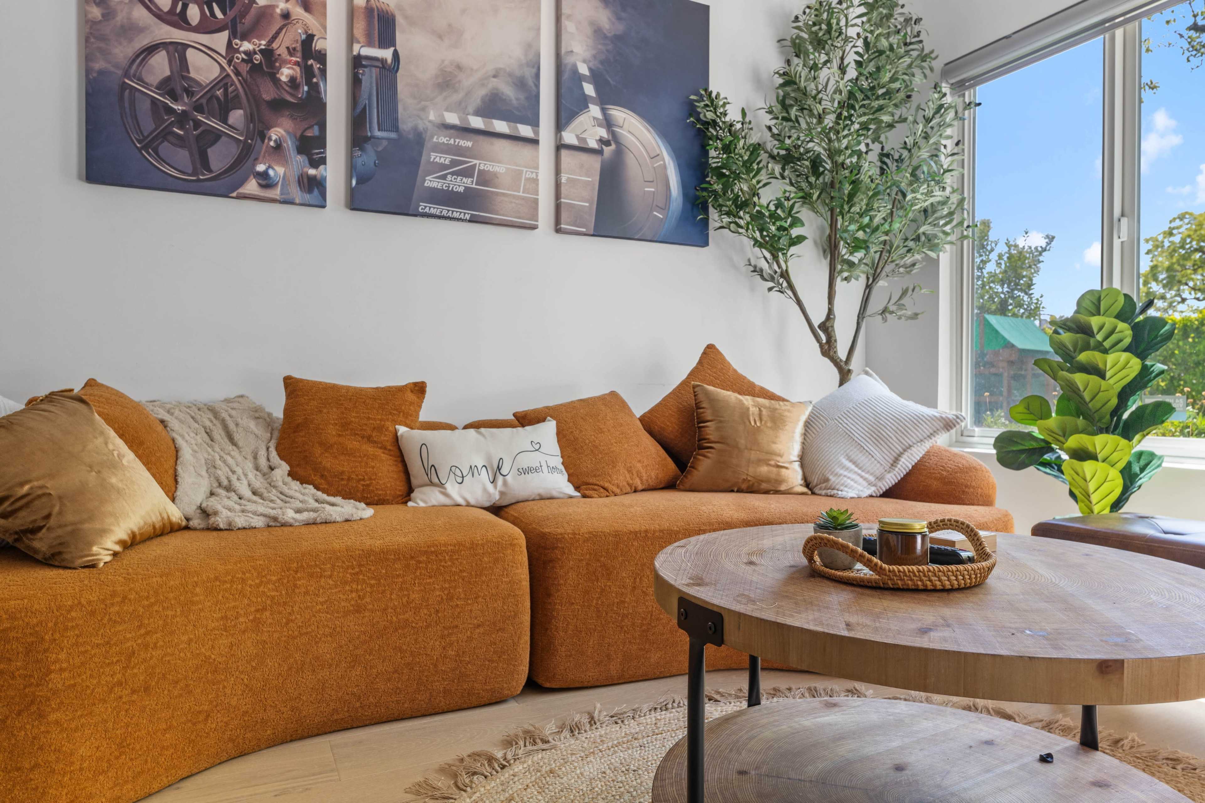 The image shows a cozy living room featuring an orange sectional sofa, a wooden coffee table, and decorative plants, with framed photographs on the wall.