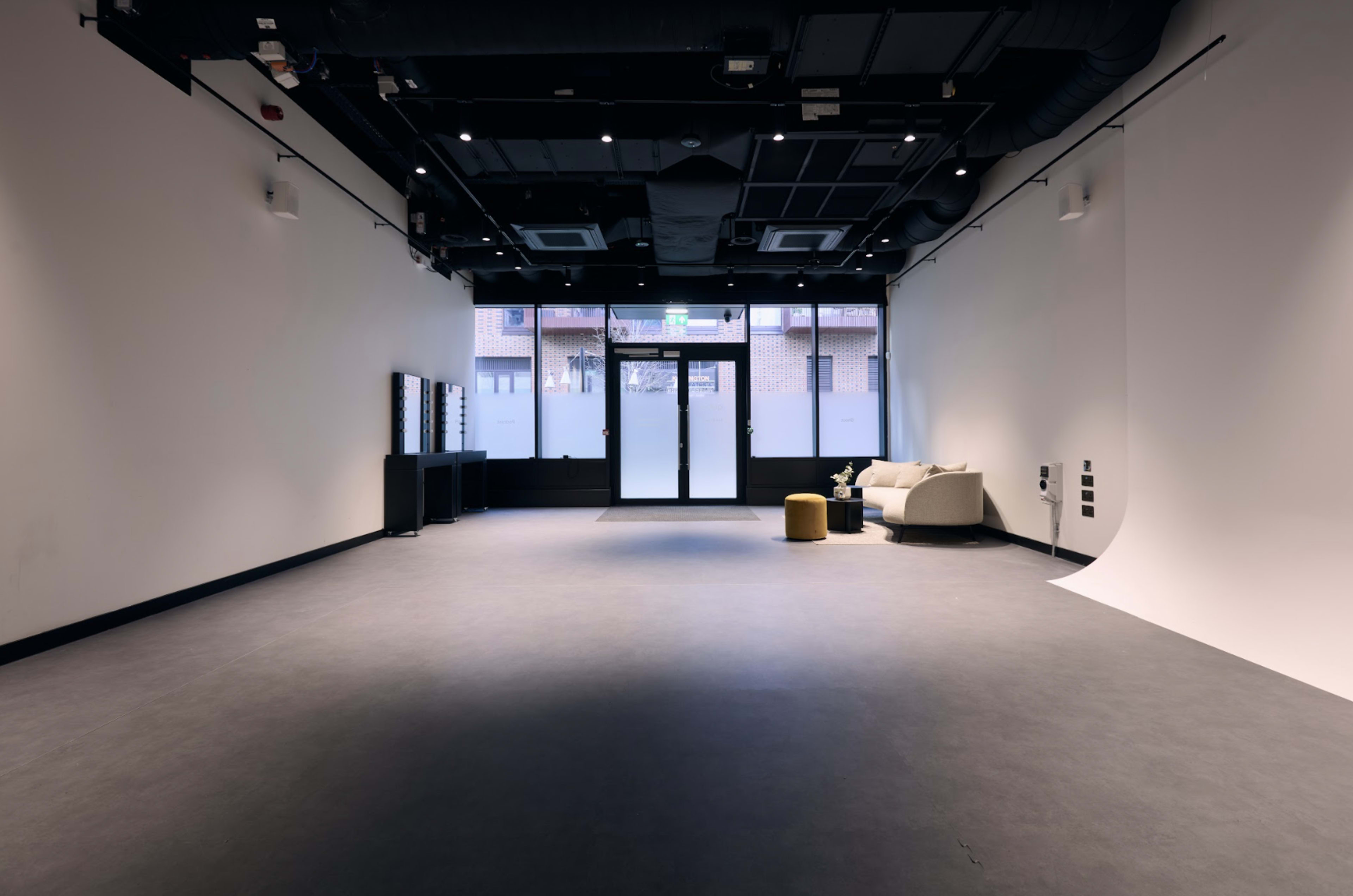 The image shows a spacious, empty room with gray flooring, a beige couch, a small table, and a large glass door that opens to an outdoor area.