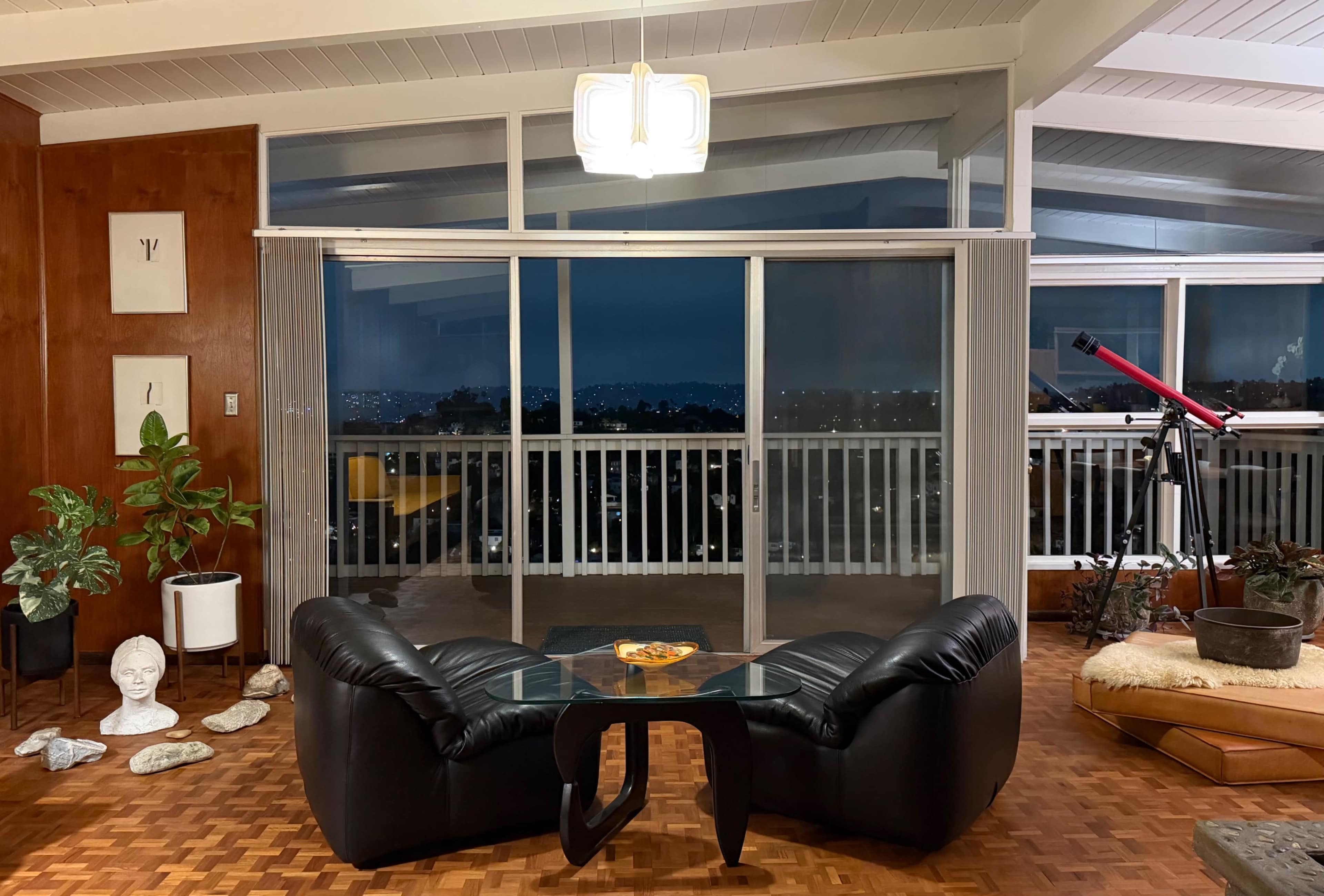 The image shows a modern living room with black leather chairs, a glass coffee table, large sliding glass doors revealing a city view, and a telescope positioned near the window.