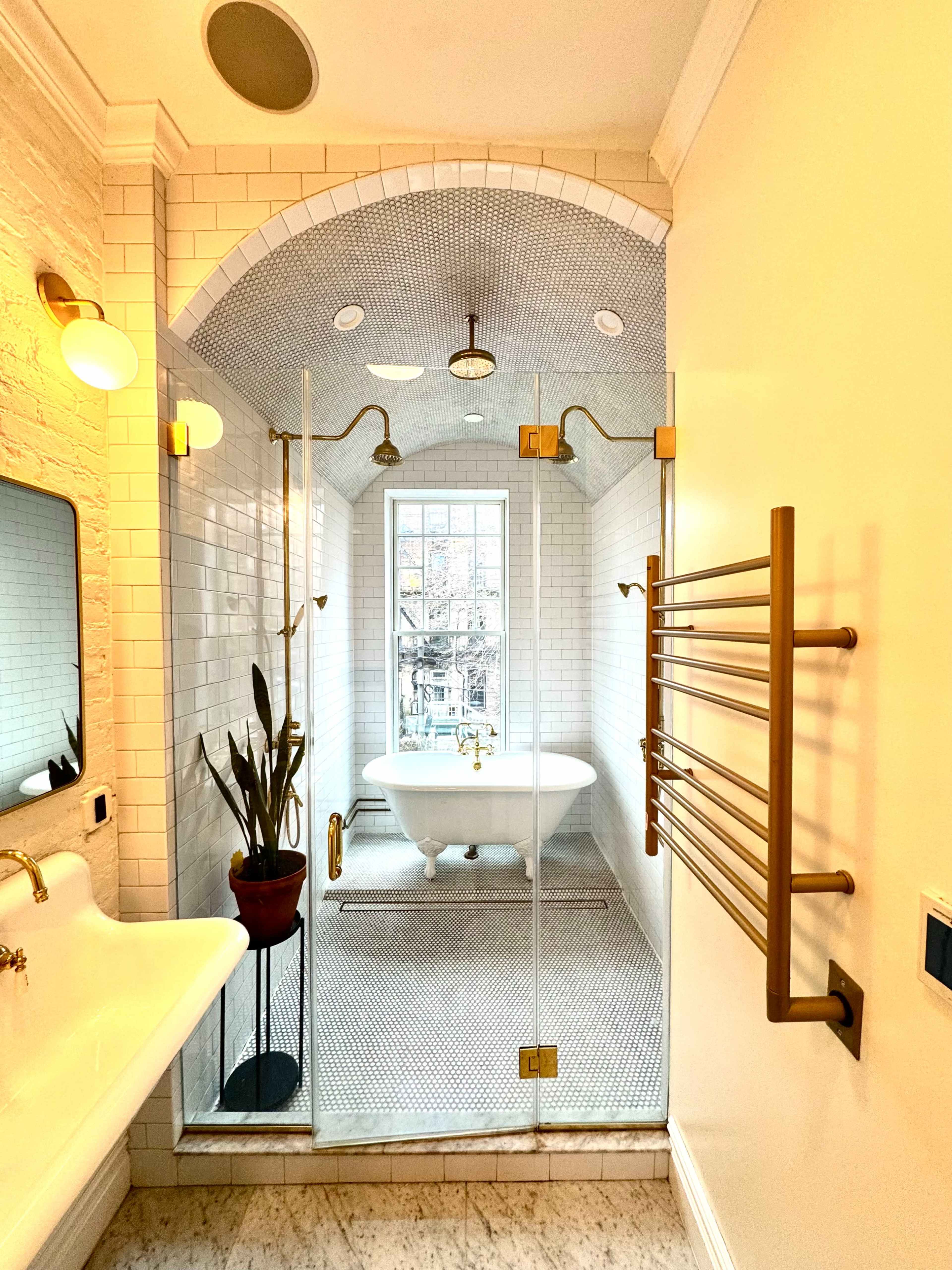 The image shows a modern bathroom featuring a glass shower enclosure, a freestanding bathtub, a wall-mounted towel rack, and a large window providing natural light.