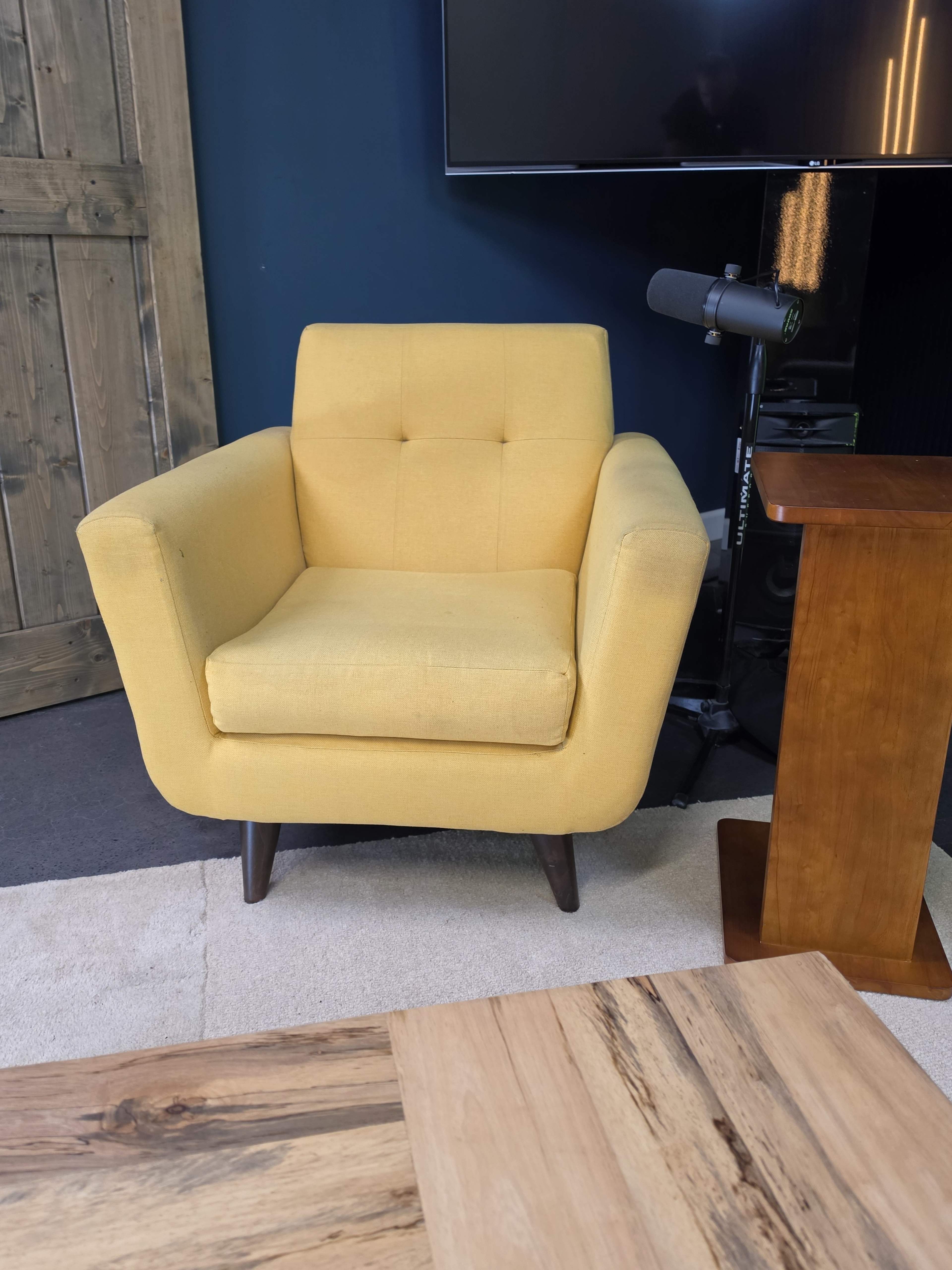 The image shows a yellow upholstered armchair with wooden legs positioned near a wooden table and a wall-mounted TV.
