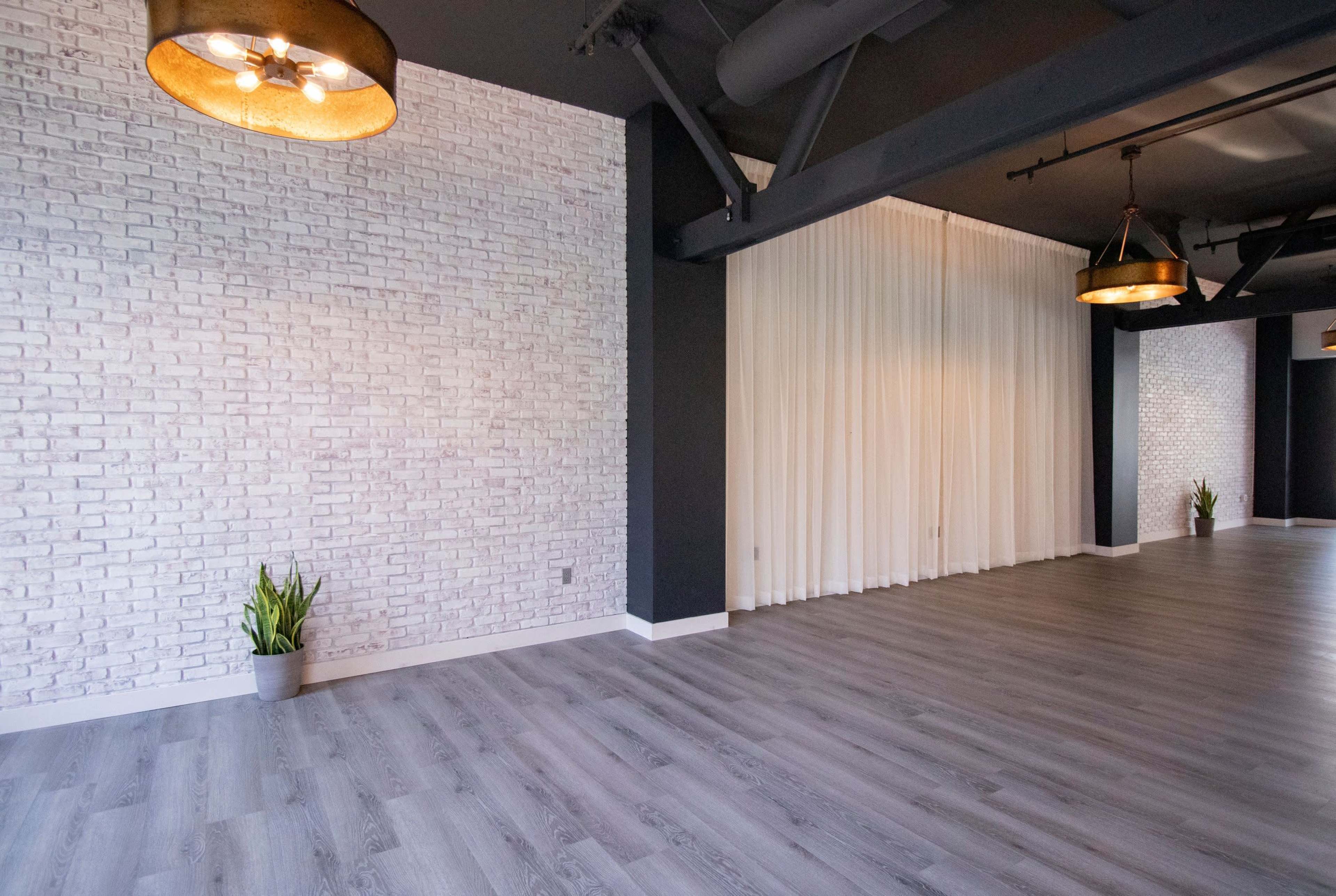 The image shows a room with a light gray wooden floor, white brick walls, sheer curtains on one side, and industrial-style pendant lights hanging from the ceiling.