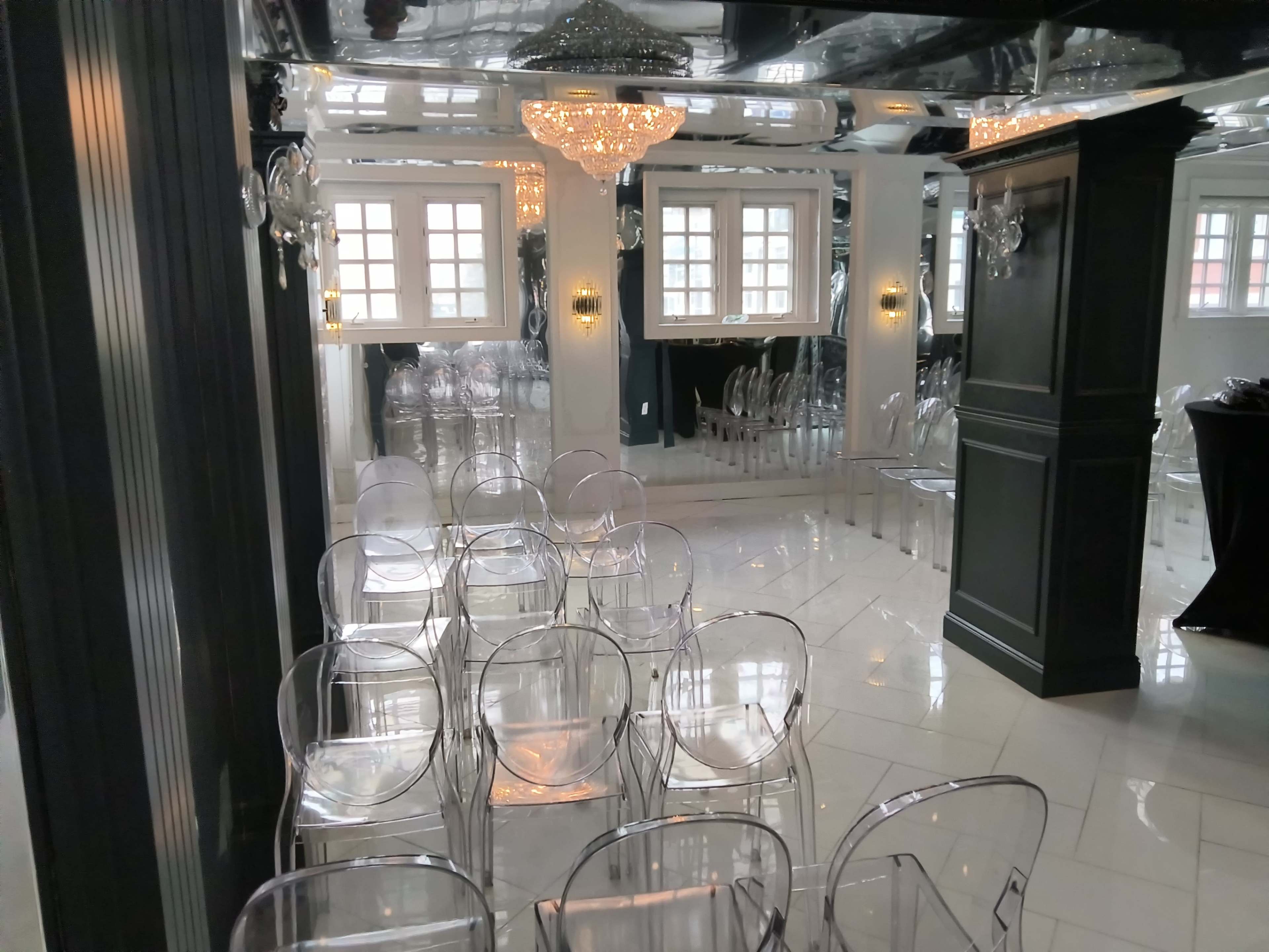 The image shows a modern event space with clear acrylic chairs arranged in rows, and large windows reflecting light from a chandelier above.